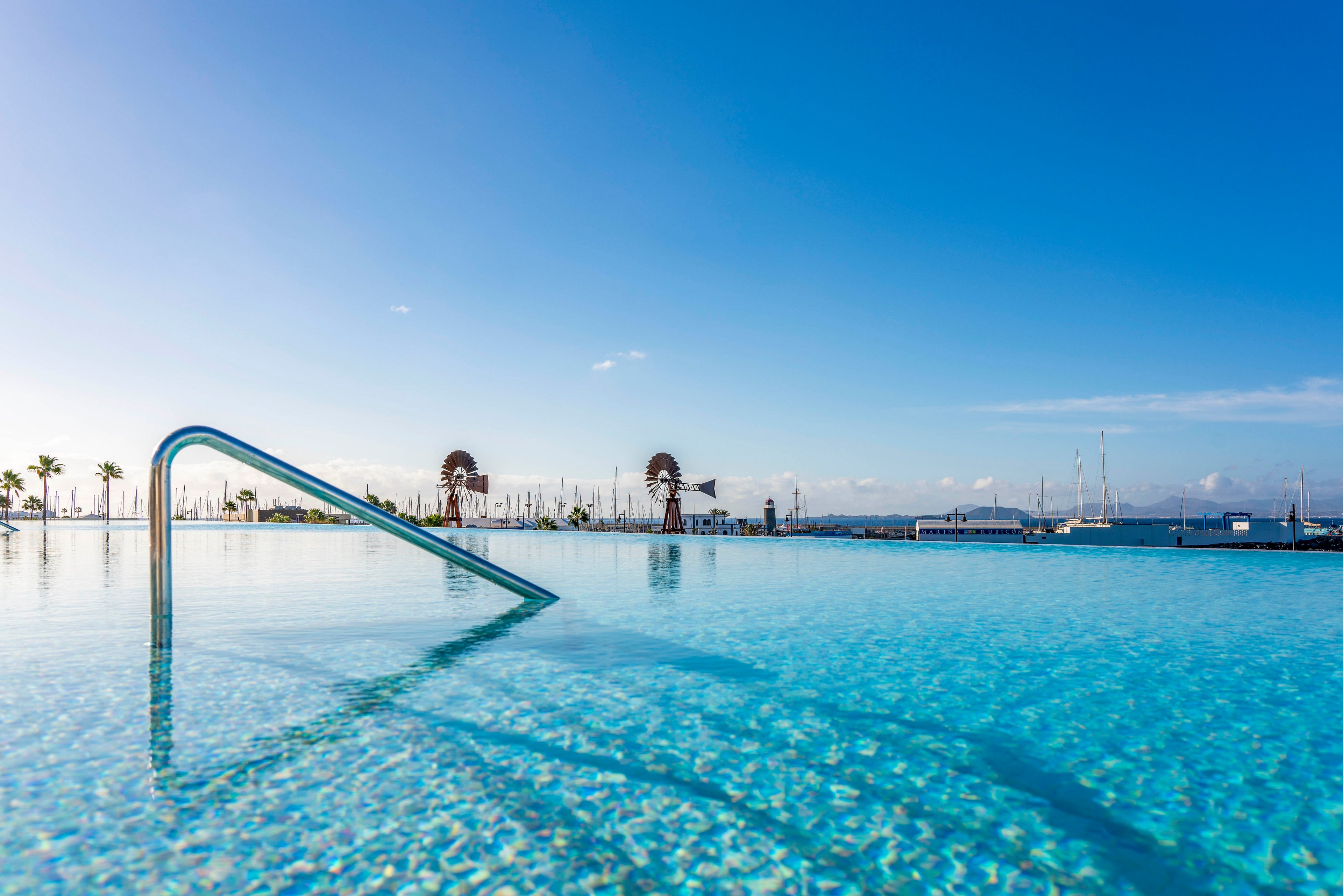 A clear blue infinity pool stretches toward the horizon under a bright, cloudless sky. A metal handrail emerges from the shallow water, leading the eye toward the distant waterfront. In the background, sailboat masts and palm trees line the marina, suggesting a coastal resort setting. The calm water and soft light create a tranquil, vacation-like atmosphere.