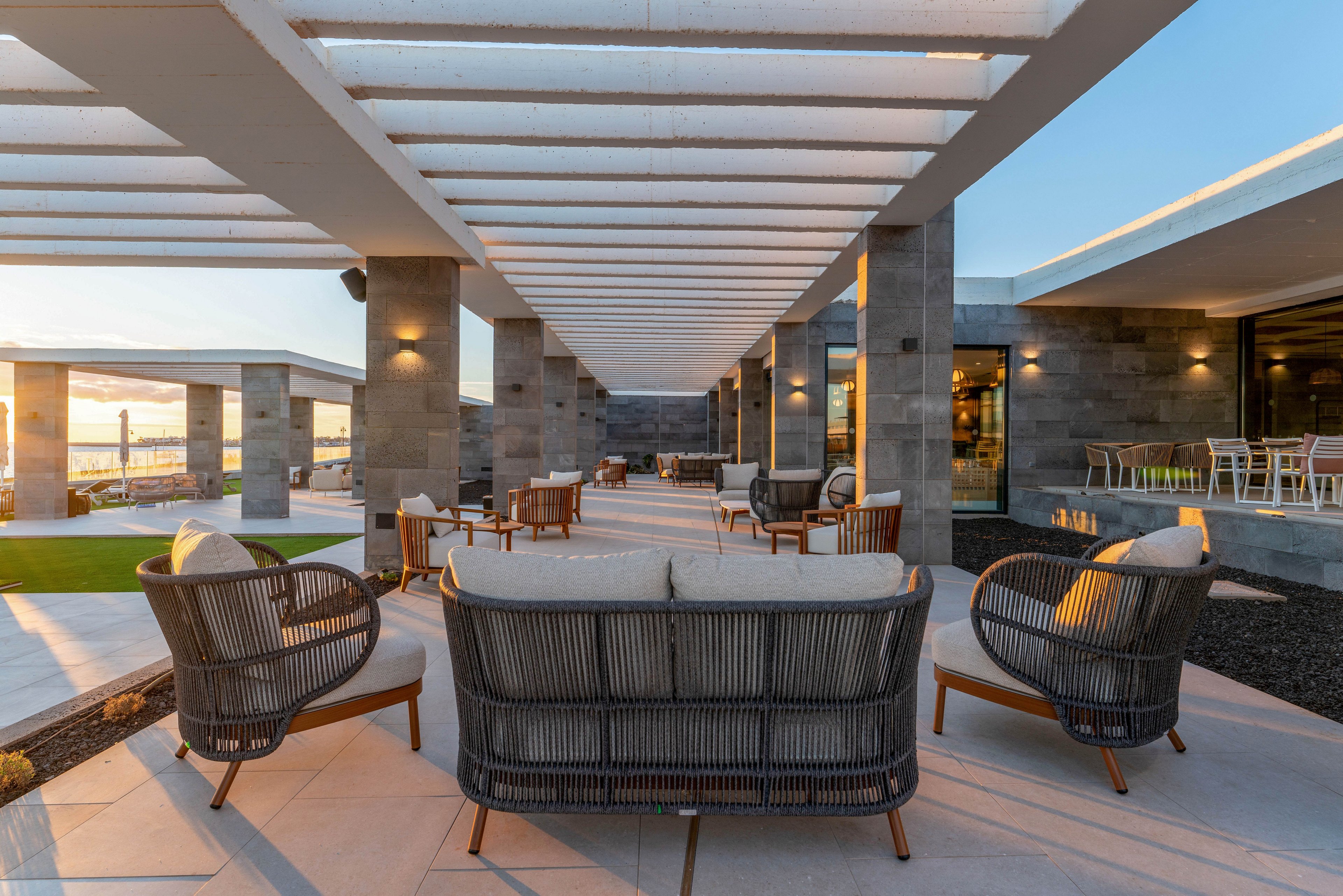 Contemporary outdoor lounge area with woven armchairs and low tables is arranged beneath a sleek pergola structure. The setting appears to be part of a modern resort or hotel terrace with clean architectural lines and stone columns. Warm evening light and visible horizon create a relaxed, inviting atmosphere. Empty seating and soft cushions emphasize comfort and leisure in an upscale environment.