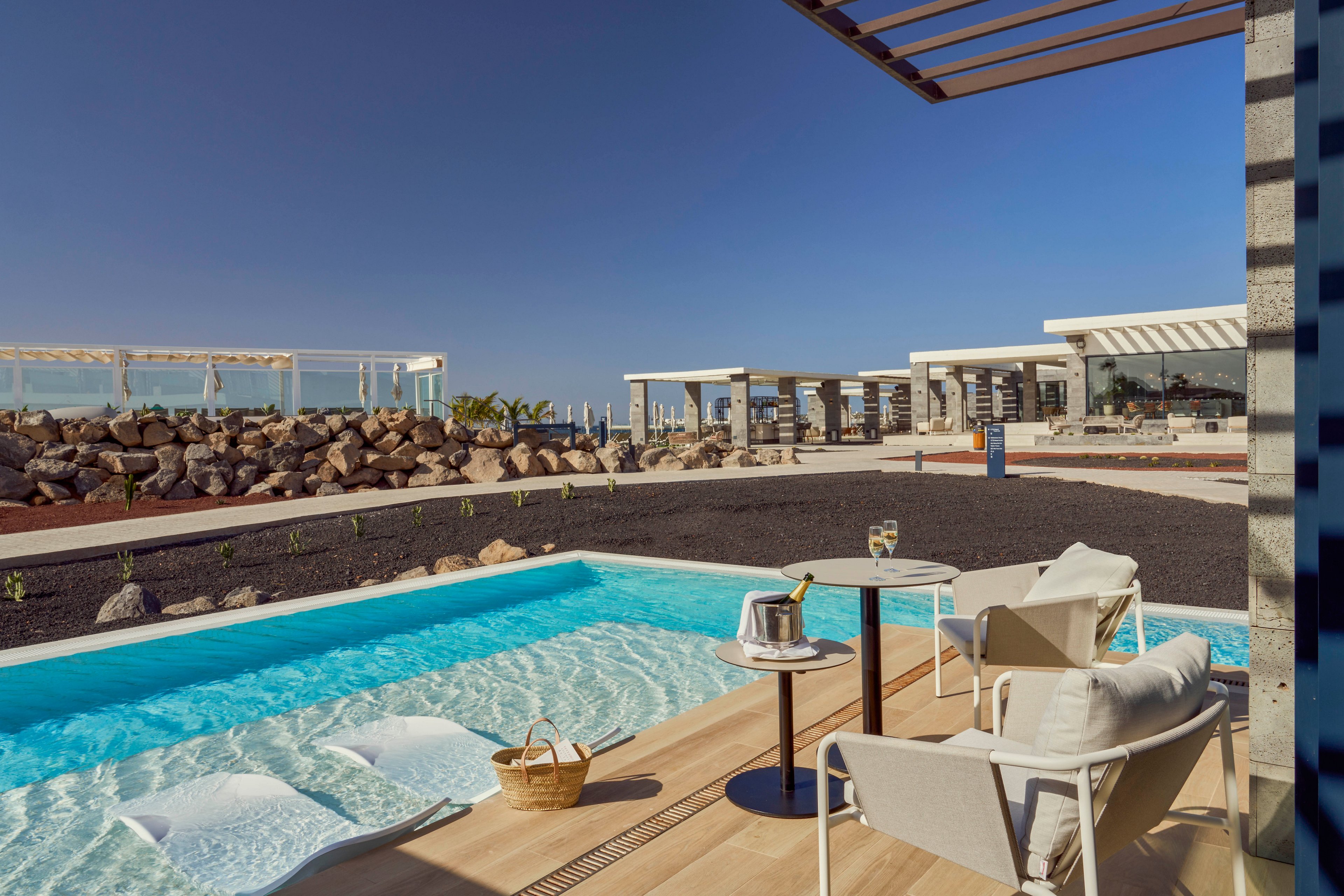 A modern resort terrace features a small private pool with submerged loungers beside a rocky coastal landscape. Two white chairs and a small table with drinks and an ice bucket create an inviting relaxation area. Contemporary white structures and pergolas line the background under a clear blue sky. The scene evokes a tranquil, upscale vacation atmosphere with strong contrasts between turquoise water, dark volcanic ground, and light architecture.
