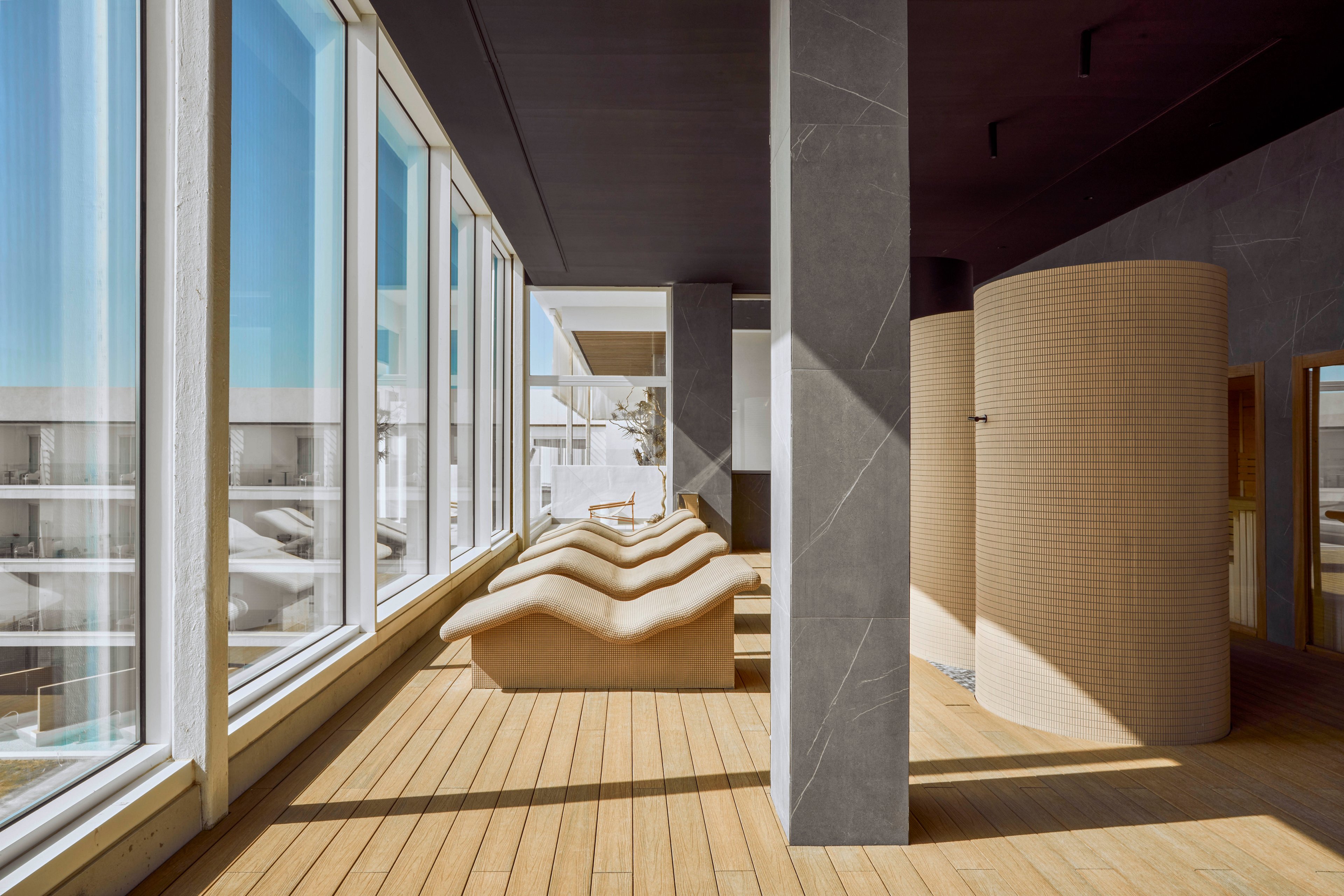 A contemporary spa relaxation area features a row of sculpted beige loungers. Large floor-to-ceiling windows flood the wooden deck floor with natural light and shadows. Curved tiled partitions and dark ceiling panels add a minimalist architectural touch. The space appears calm and serene, designed for rest and wellness.