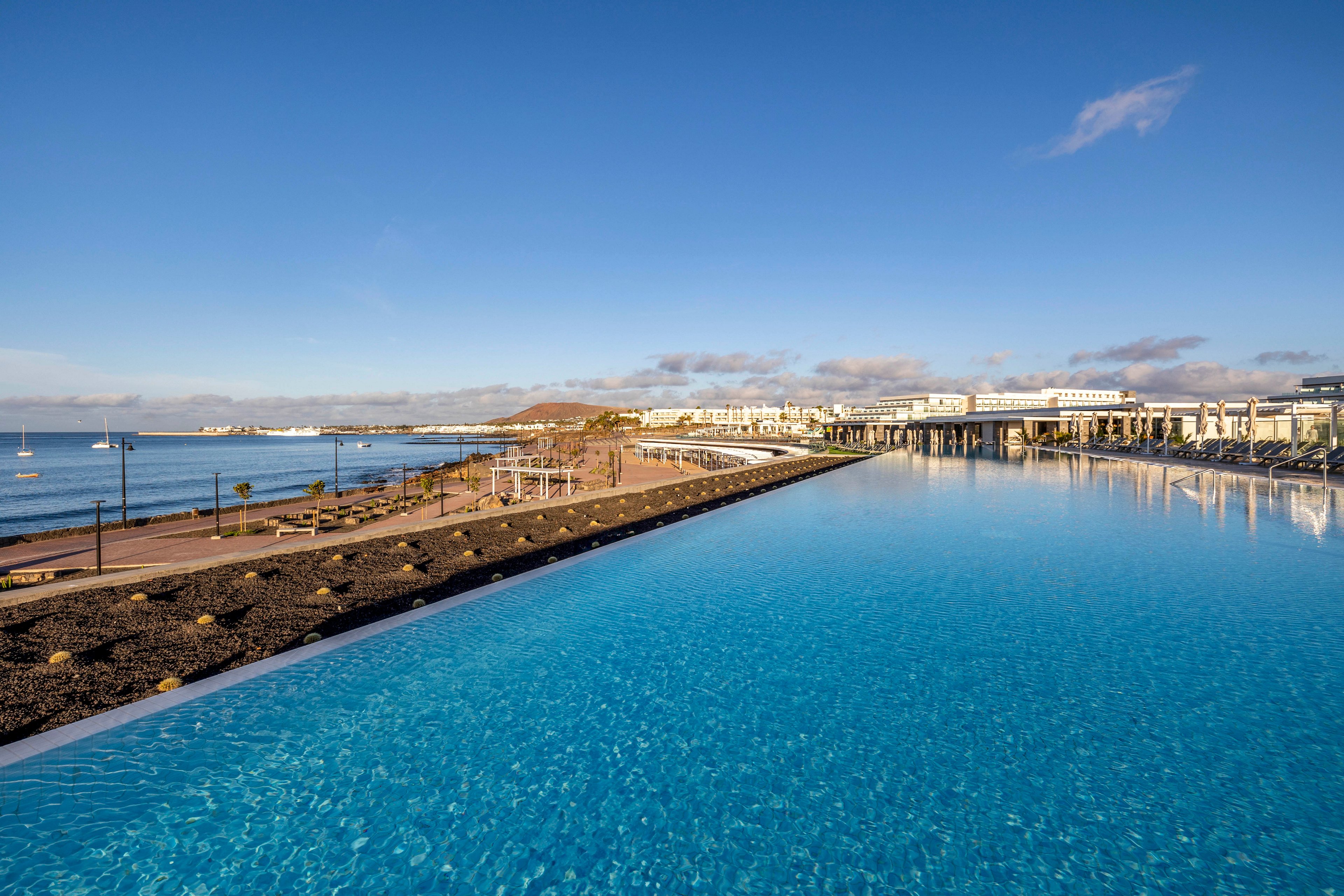A large infinity pool stretches toward the horizon beside a coastal resort. Modern white buildings line the waterfront under a clear blue sky. The calm sea and distant hills create a serene backdrop with a few small boats visible offshore. Warm sunlight enhances the bright blue water and relaxed vacation atmosphere.
