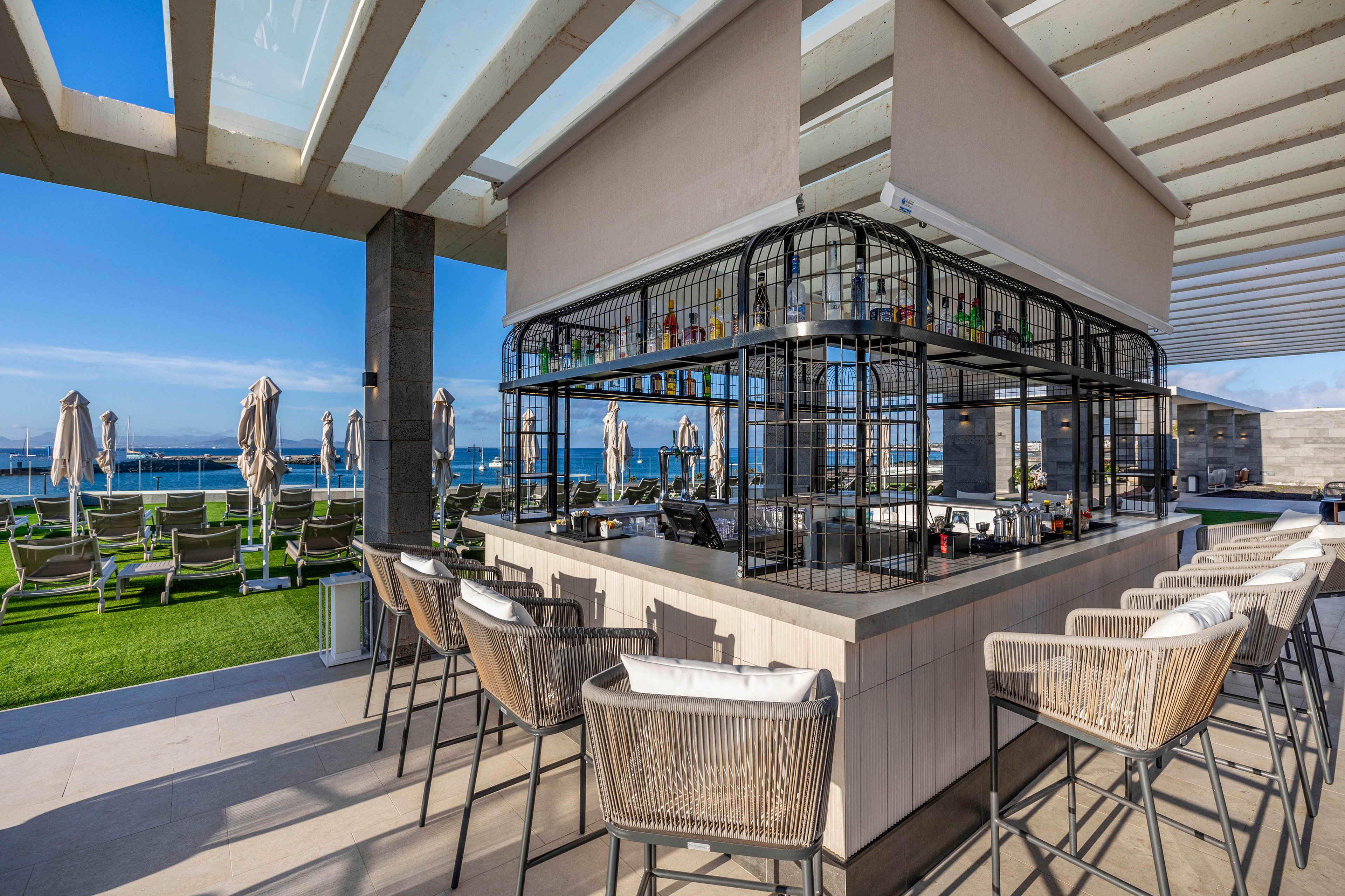 Contemporary open-air bar with high stools sits under a pergola near the ocean. The central bar island features a metal-framed shelving unit displaying various bottles. Sun loungers and closed umbrellas are arranged on a grassy area facing the sea in the background. Bright daylight and clear blue sky create a relaxed, vacation atmosphere.