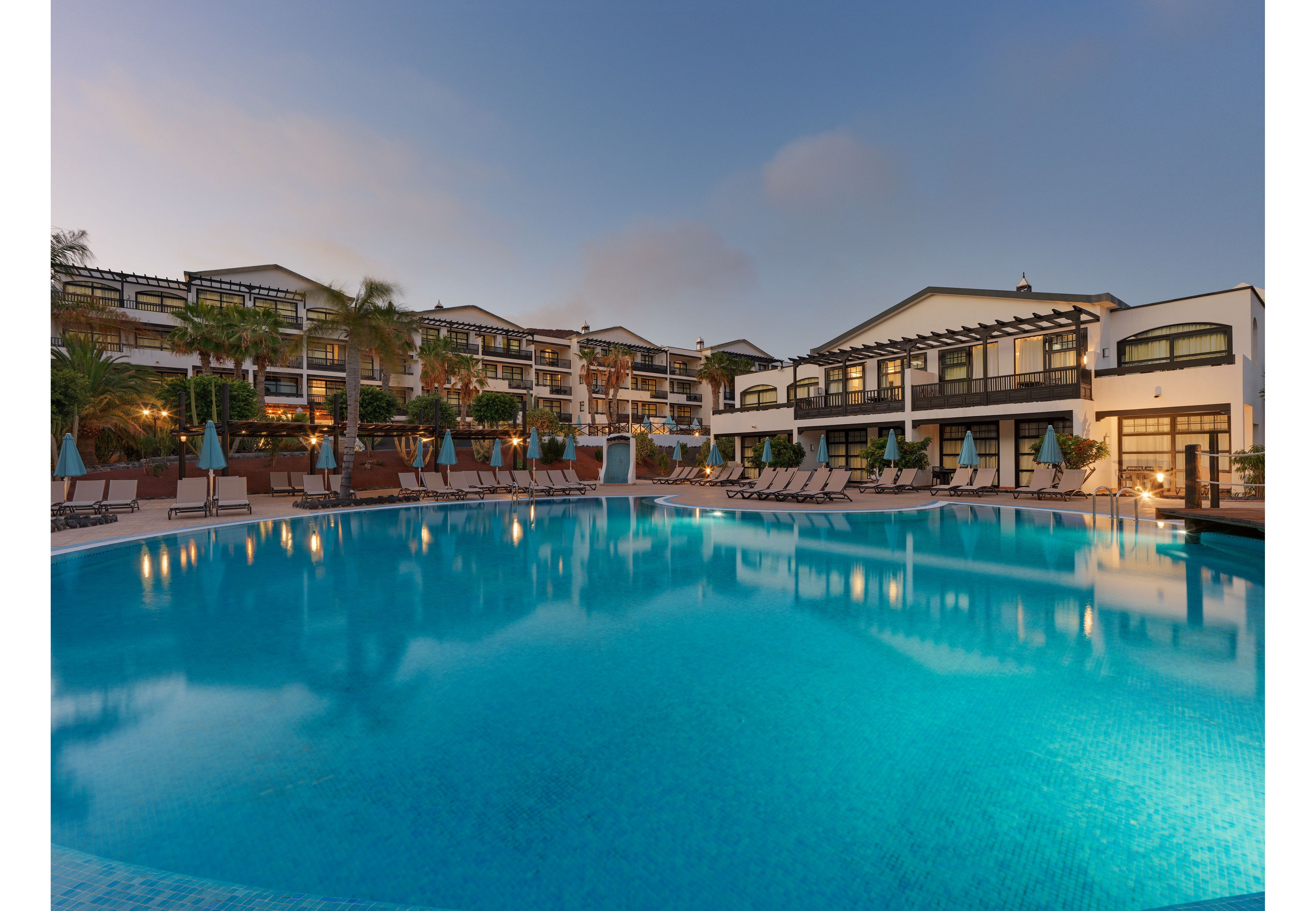 A large, tranquil swimming pool reflects the lights of a modern resort complex at dusk. Multi-story buildings with balconies and terraces surround the pool, framed by palm trees and landscaped walkways. Lounge chairs and umbrellas line the poolside, suggesting a relaxing vacation atmosphere. The sky transitions from soft blue to warm tones, enhancing the serene and inviting mood of the scene.