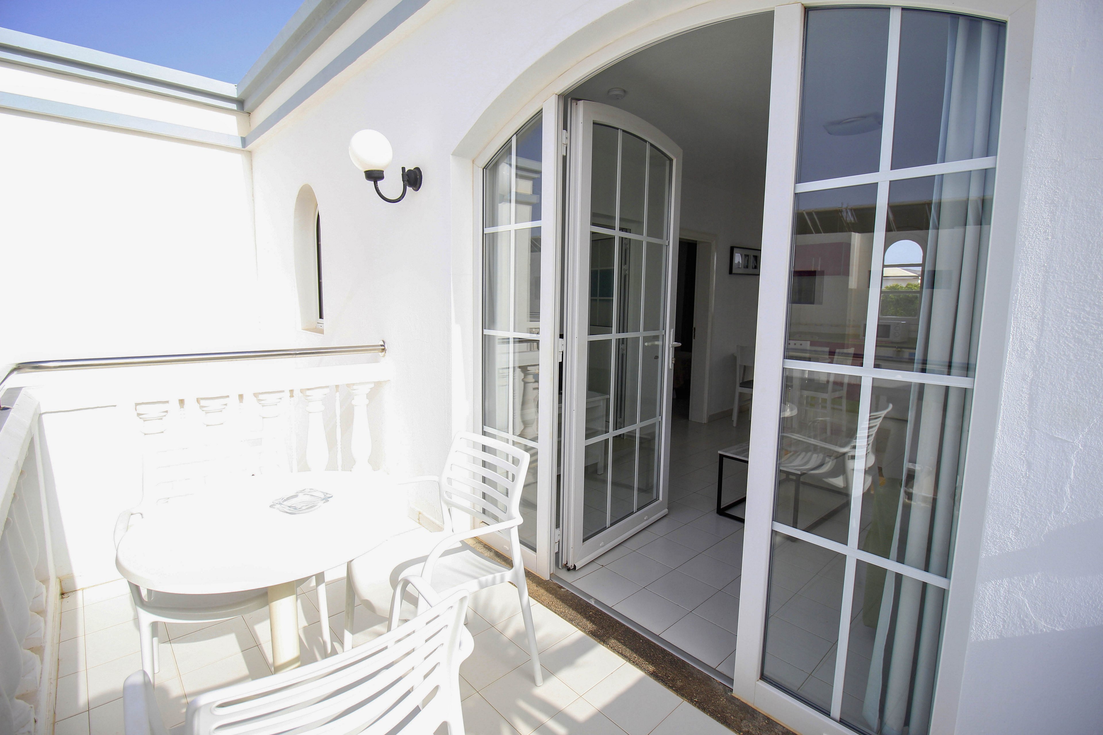 A small white balcony features a round table and several chairs in bright daylight. Large glass French doors open from the balcony into a modern interior room. The space is dominated by clean white walls, railings, and flooring, creating a minimalist and airy feel. A wall-mounted light fixture and arched window details add subtle architectural interest.