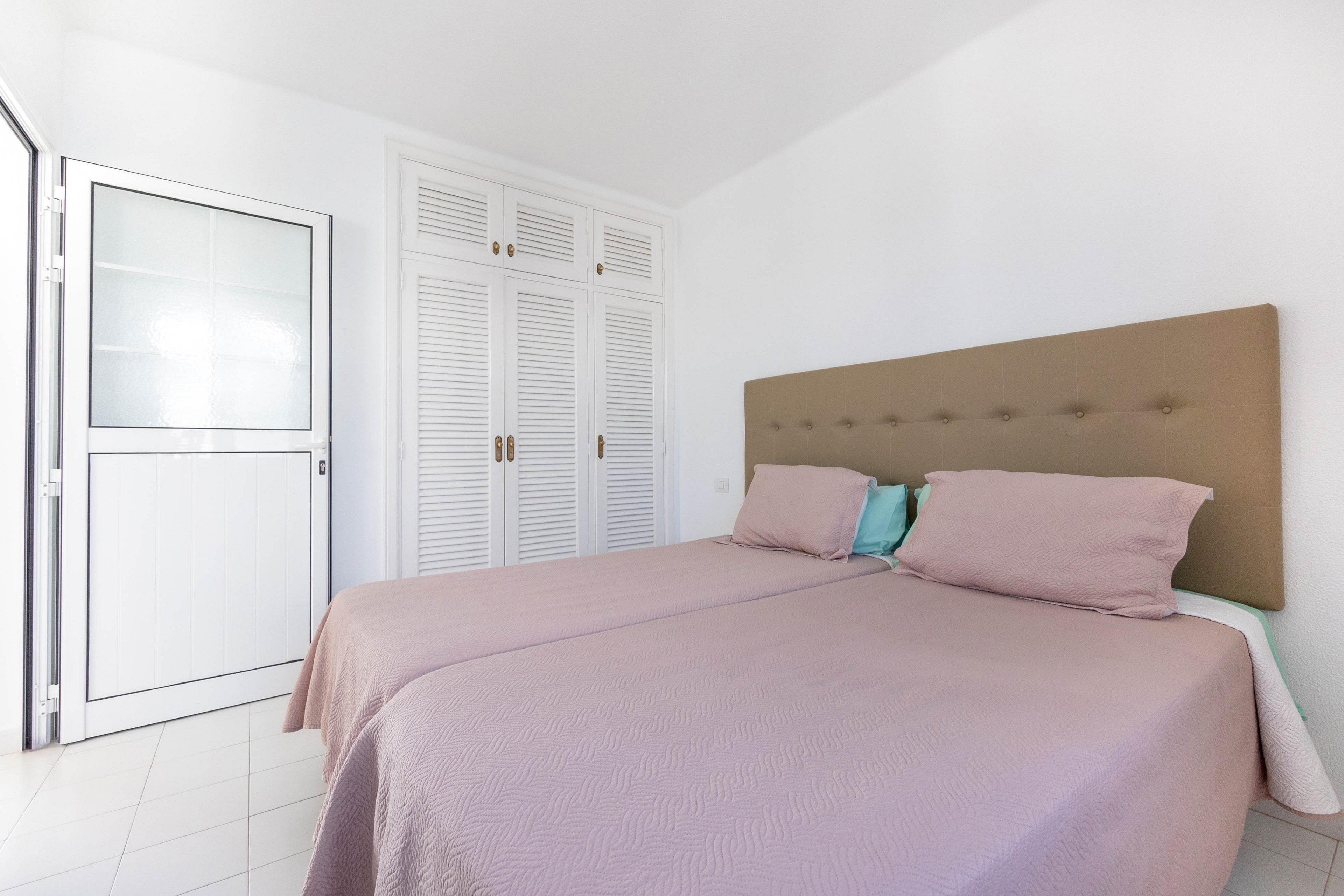 A bright, minimalist bedroom features two twin beds pushed together with a shared upholstered headboard. Soft mauve bedding and pillows add a subtle touch of color against the white walls and floor. Built-in white louvered wardrobes line the wall beside a white door with frosted glass panels. The room appears clean, airy, and uncluttered, emphasizing simplicity and calm.