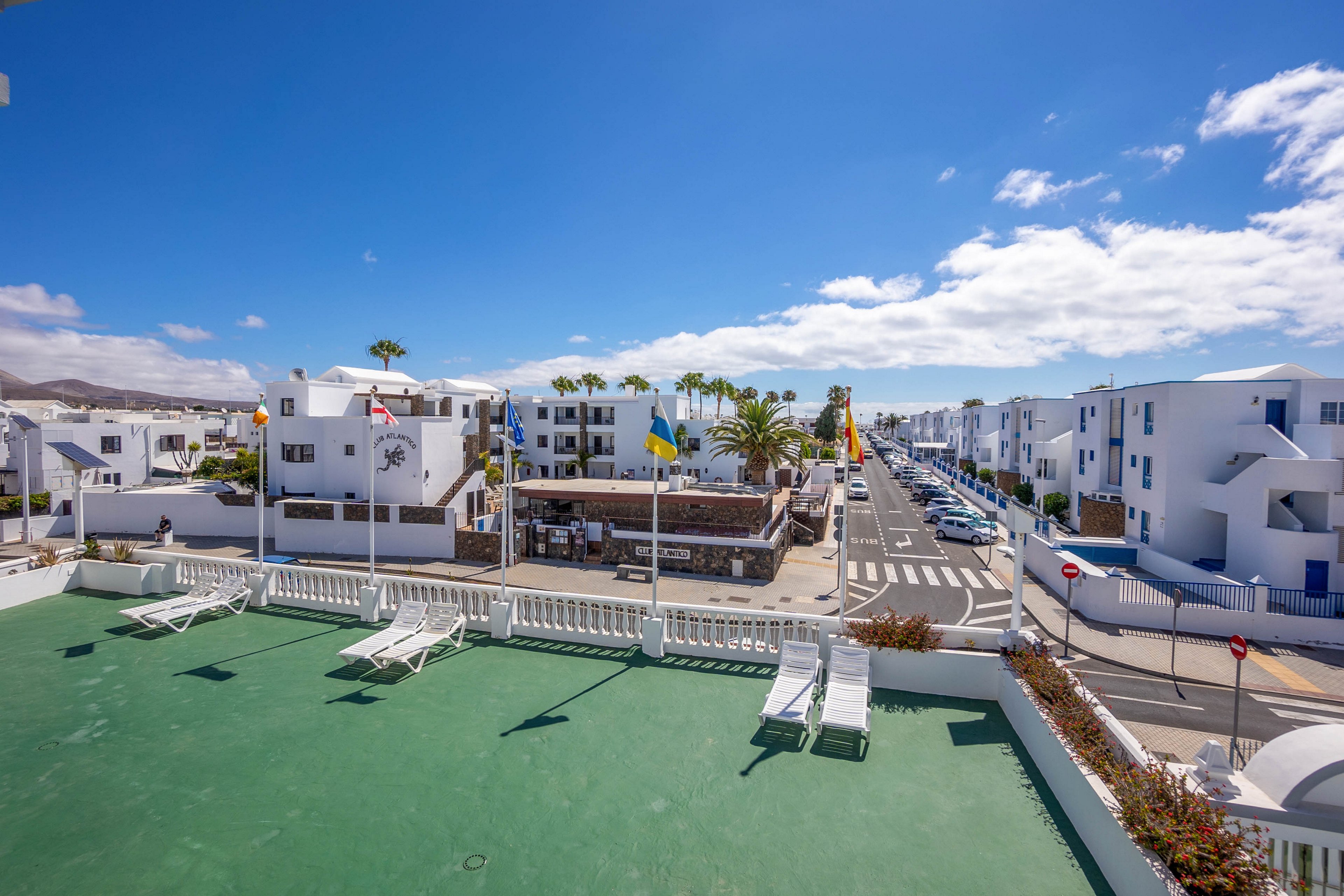 A bright coastal town is viewed from an elevated rooftop terrace with several white sun loungers. White low-rise apartment buildings line both sides of a straight street that stretches toward the horizon. Flags flutter above a small central building, and parked cars are visible along the road. The sky is clear and blue with scattered clouds, creating a relaxed holiday atmosphere.