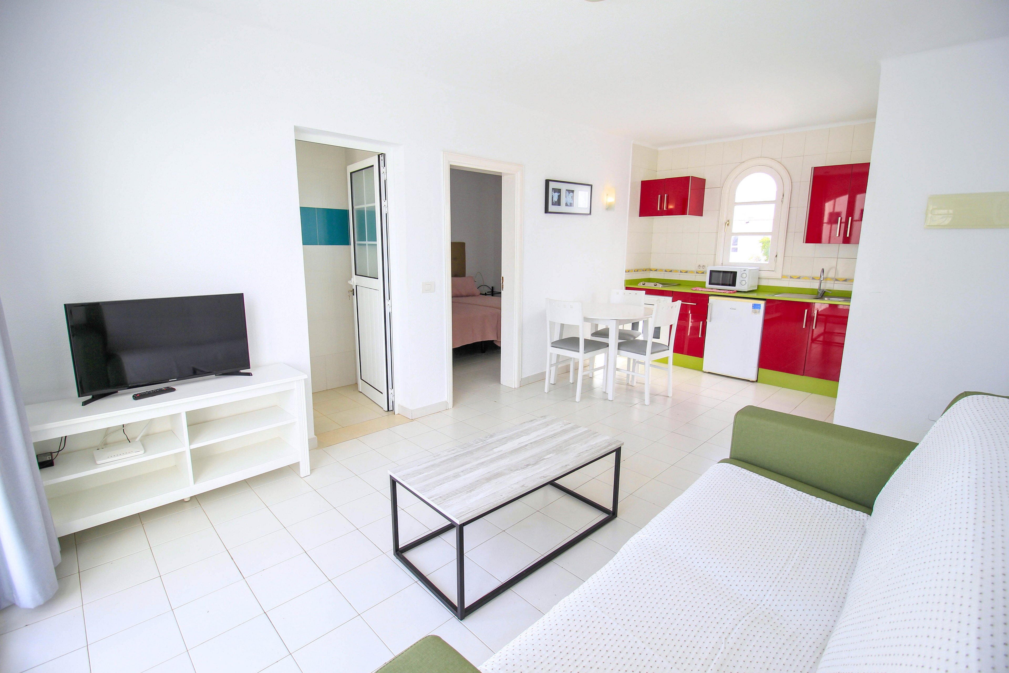 A bright modern studio apartment features a small living area with a green sofa, white coffee table, and flat-screen television on a low stand. The open-plan space extends into a compact kitchen with red cabinets, white appliances, and a dining table with four chairs. White tiled floors and walls create a clean, minimalist look throughout the room. A doorway leads to an adjacent bedroom, adding depth to the interior scene.