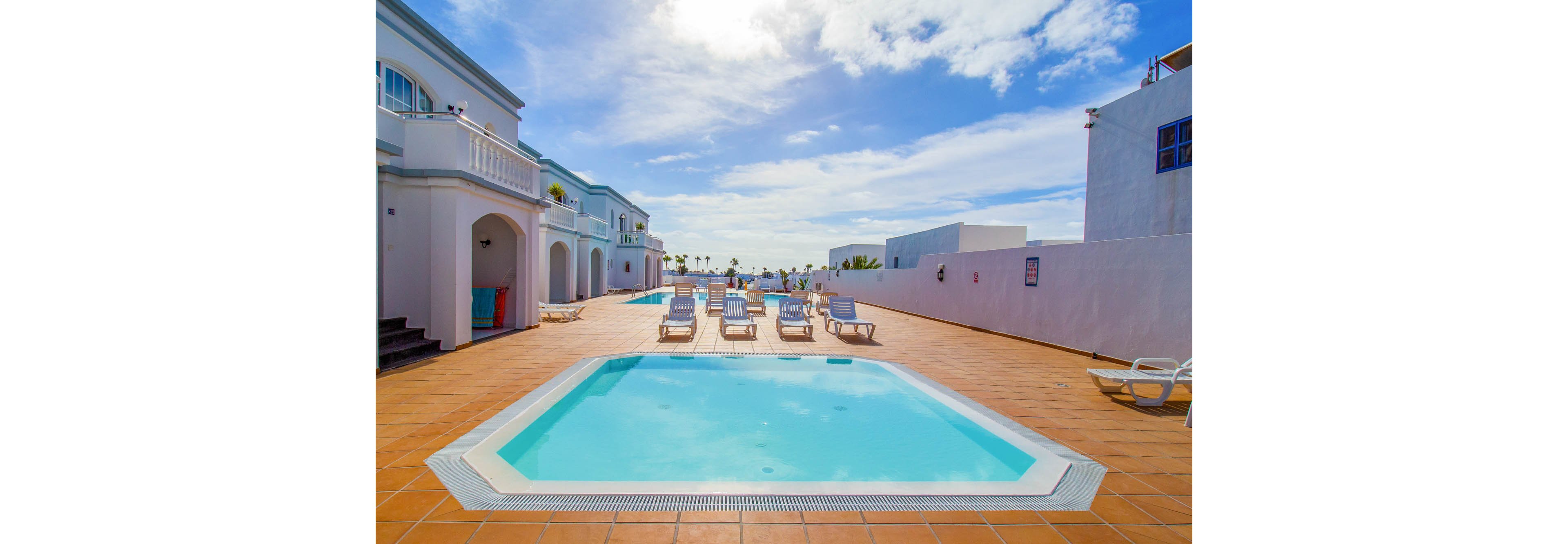 An outdoor swimming pool sits in the center of a sunlit resort courtyard. White lounge chairs line the poolside area on a wide terracotta-tiled deck. White buildings with balconies and windows surround the space under a bright blue sky with scattered clouds. The scene suggests a warm, relaxing vacation atmosphere at a coastal or island destination.