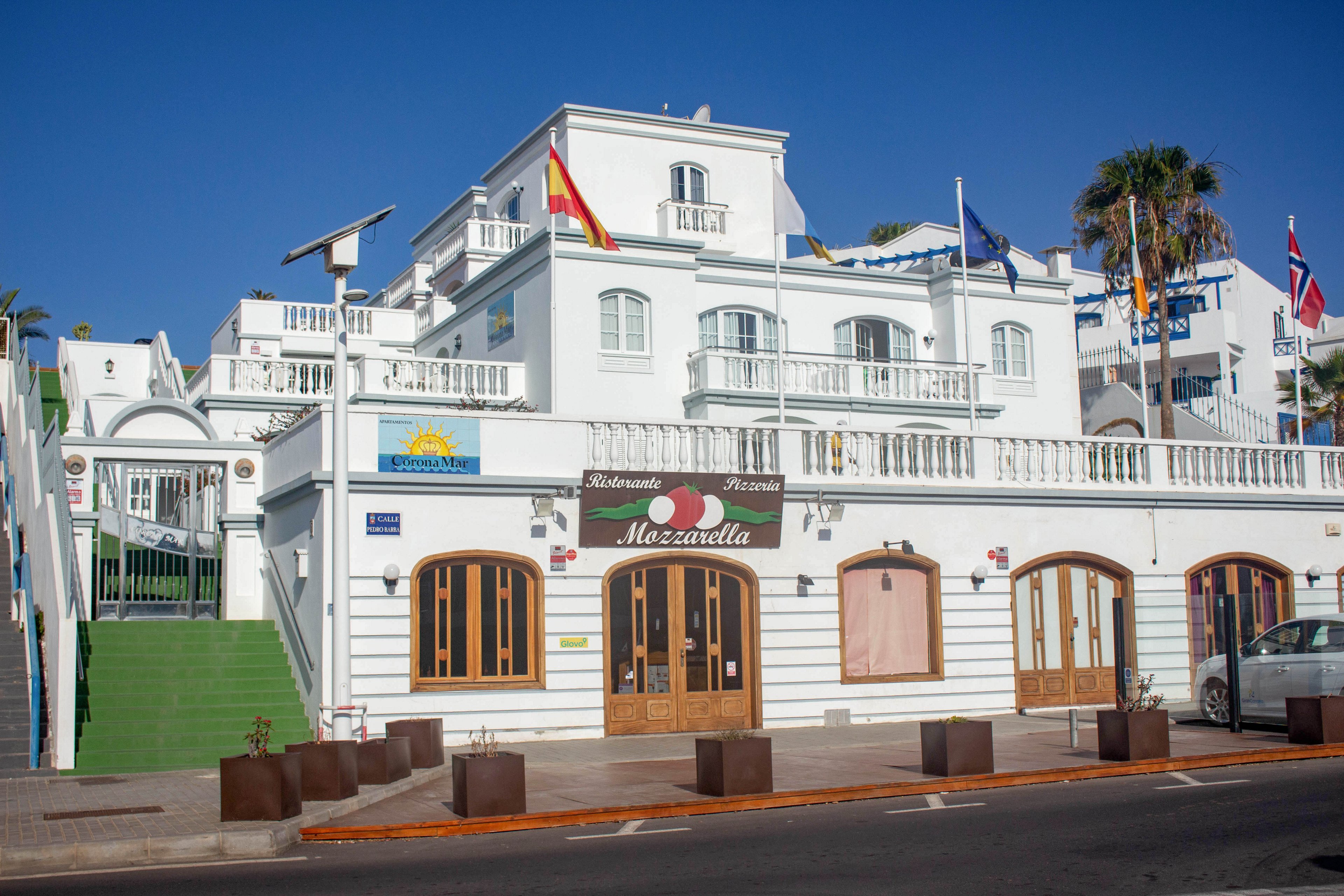 A white multi-story hotel building features arched wooden doors and balconies under a clear blue sky. The ground floor hosts a restaurant named "Mozzarella" with a red sign and heart graphic. Several national flags, including the Spanish flag, are displayed along the rooftop and terraces. A green stairway on the left leads up to the upper level, and a parked car is partially visible on the right side of the street.