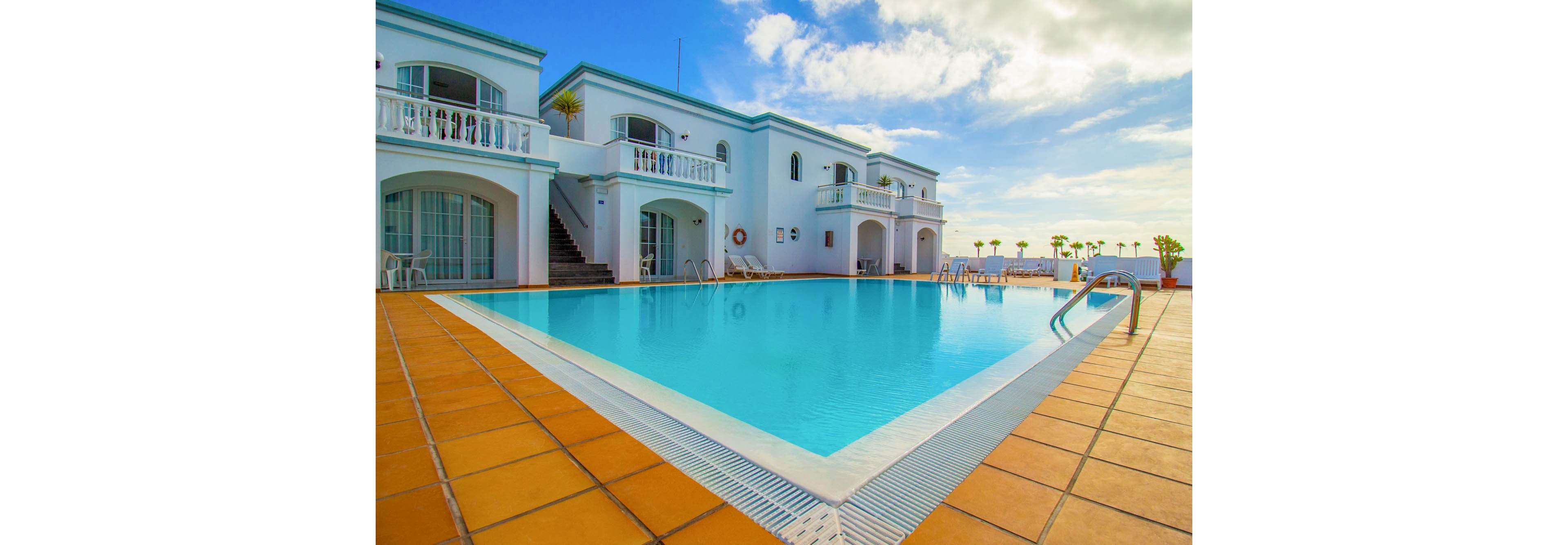 A large turquoise swimming pool sits at the center of a white Mediterranean-style resort. Two-story villas with balconies and arched windows surround the pool area. Sun loungers and umbrellas line the far side, facing an open view toward the sea and sky. The scene is brightly lit with warm sunlight and a few scattered clouds, creating a relaxed vacation atmosphere.