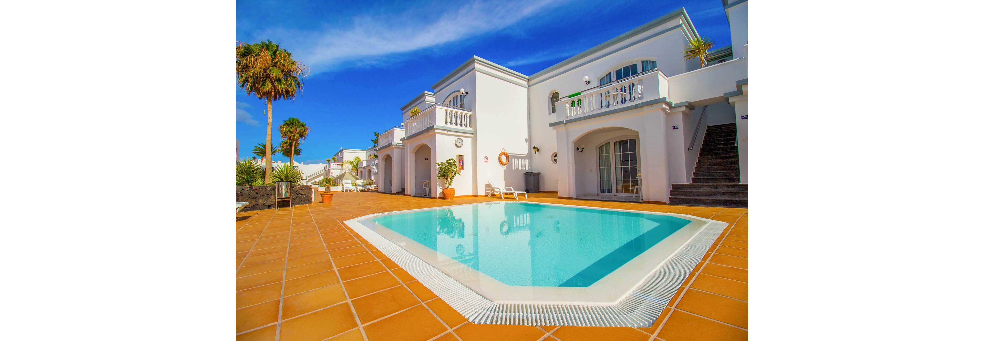 A clear turquoise swimming pool sits at the center of a sunny resort courtyard. White two-story villas with balconies and arched details surround the pool area. Terracotta tiles frame the water, with a few lounge chairs placed near the edge. Palm trees and bright blue sky enhance the warm, tropical vacation atmosphere.