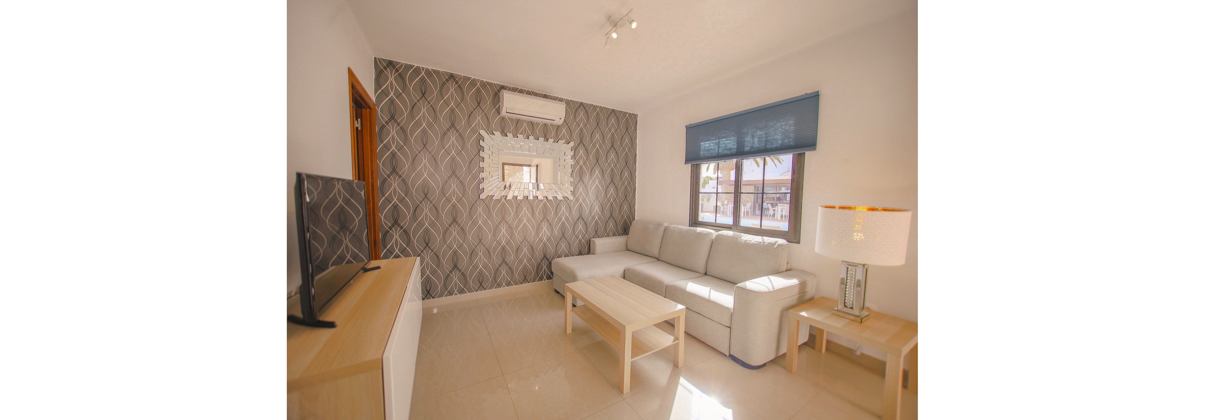 A contemporary living room features a light gray sectional sofa and simple wooden coffee table. The space includes a patterned accent wall with a decorative mirror and a wall-mounted air conditioner above it. A flat-screen television sits on a low media console opposite the sofa. Large windows with a dark roller shade let in natural light, while a table lamp on a side table adds additional illumination.