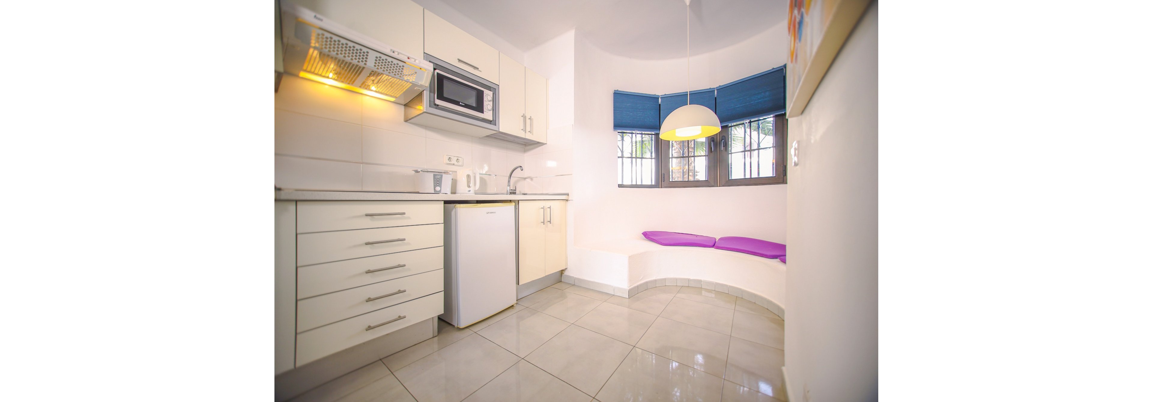 A compact modern kitchenette features white cabinets, a small refrigerator, and built-in microwave. The space is clean and minimal with glossy tiled floors and light-colored walls. A curved window nook with blue blinds and purple cushions adds a cozy seating area. A hanging pendant lamp illuminates the corner, creating a bright and inviting atmosphere.
