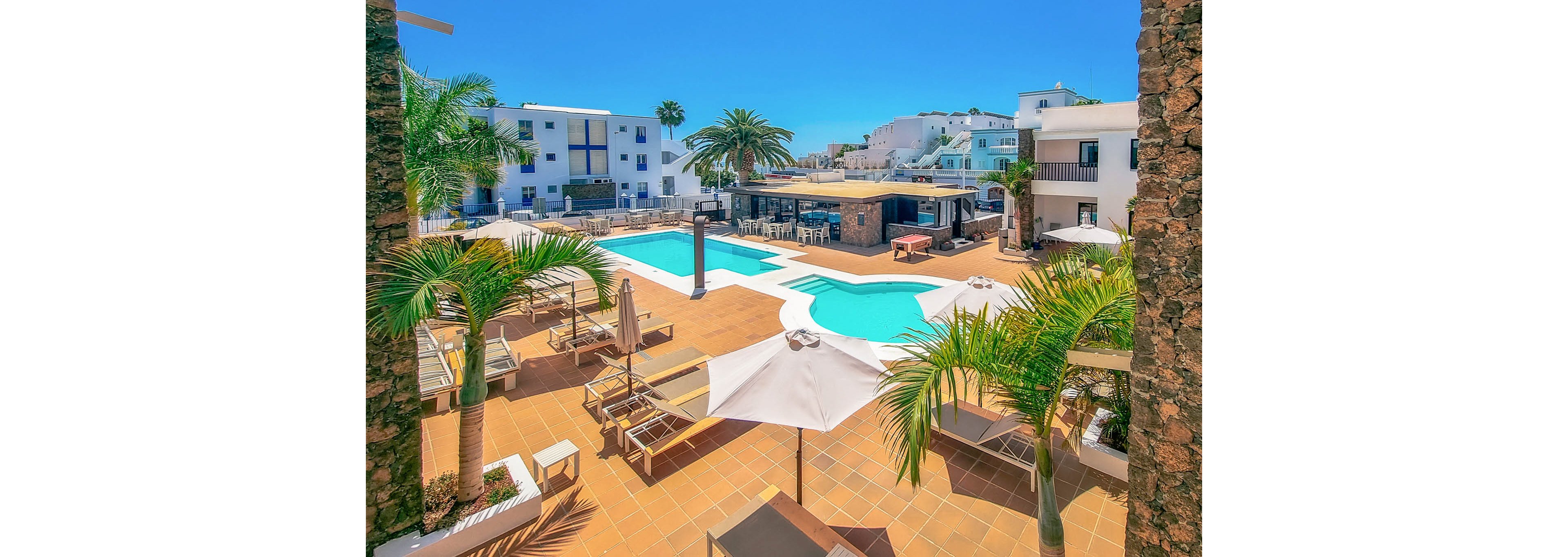 A bright resort courtyard features a large swimming pool surrounded by sun loungers and umbrellas. Palm trees and tropical plants frame the tiled terrace, creating a relaxing vacation atmosphere. A poolside bar and seating area sit at the center, with white low-rise buildings in the background. The clear blue sky and turquoise water emphasize the warm, sunny climate.