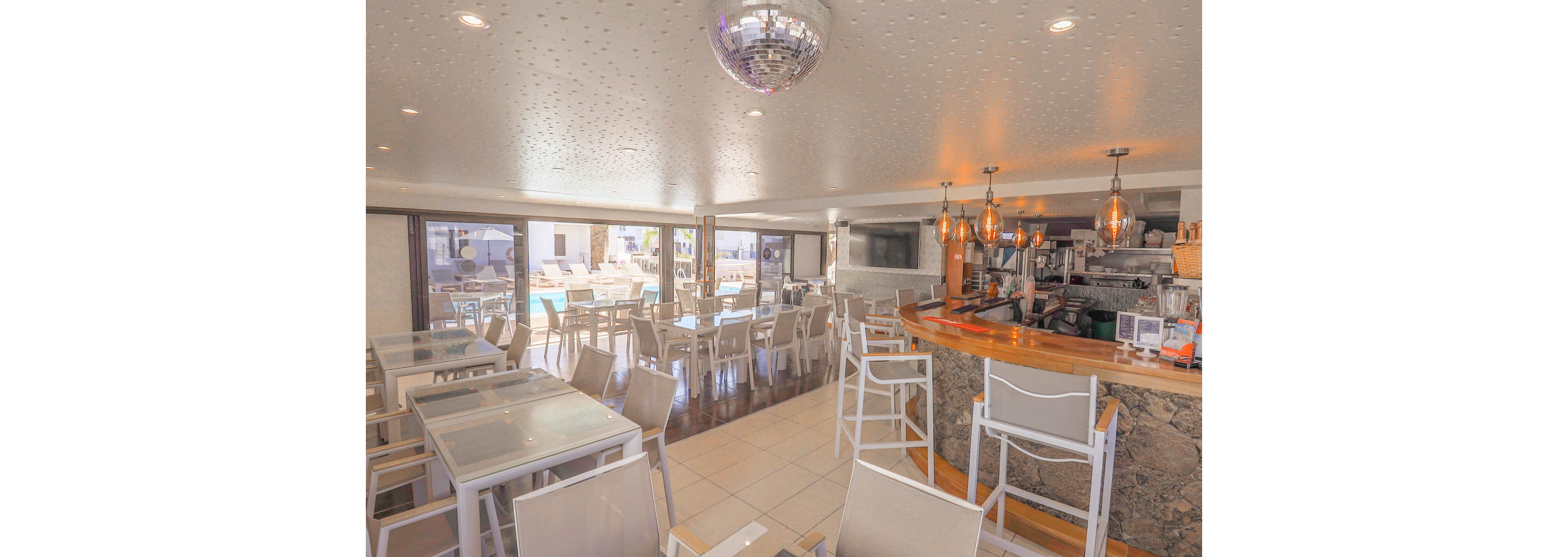 Spacious bar and restaurant area with modern tables and chairs is shown in soft daylight. A curved wooden counter with barstools faces an open kitchen and drink preparation area. Large windows and open walls reveal an outdoor poolside terrace with additional seating. Warm pendant lights and a ceiling disco ball add a relaxed, vacation atmosphere.