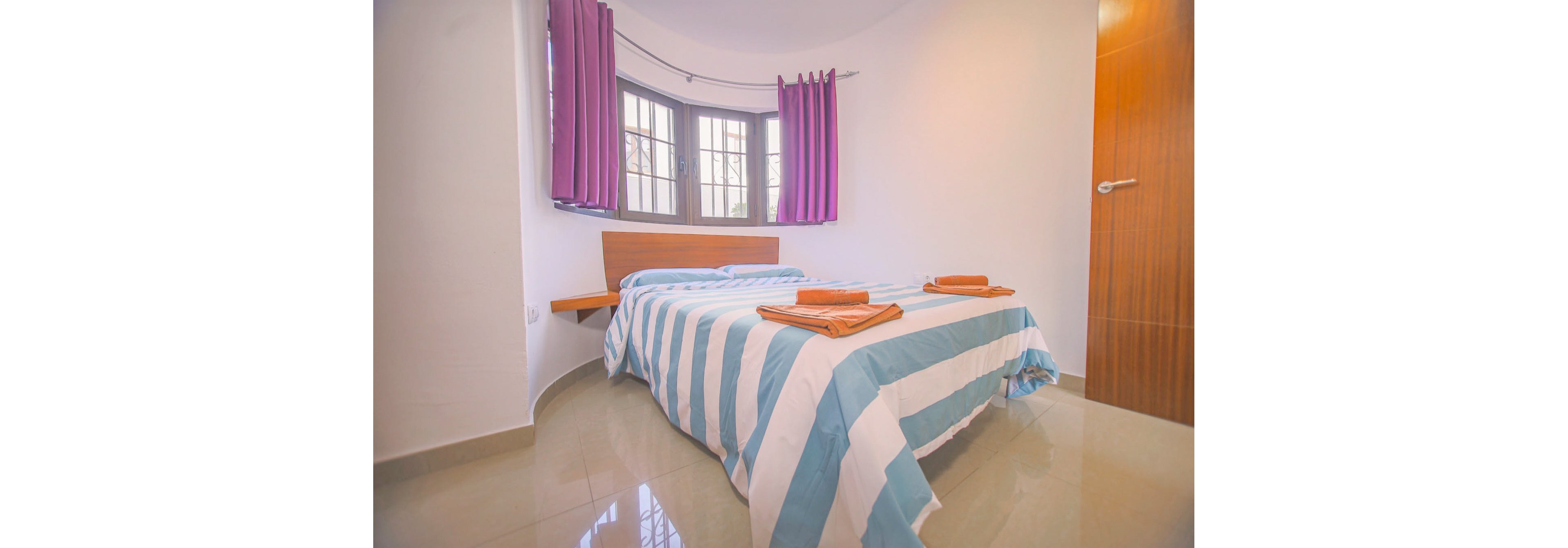 A small modern bedroom features a bed with blue and white striped bedding as the central focus. The room has glossy tiled floors and a wooden door on the right side. Large windows with multiple panes let in natural light and are framed by bold purple curtains. Folded orange towels are neatly placed on the bed, adding a contrasting accent color to the clean, minimalist space.