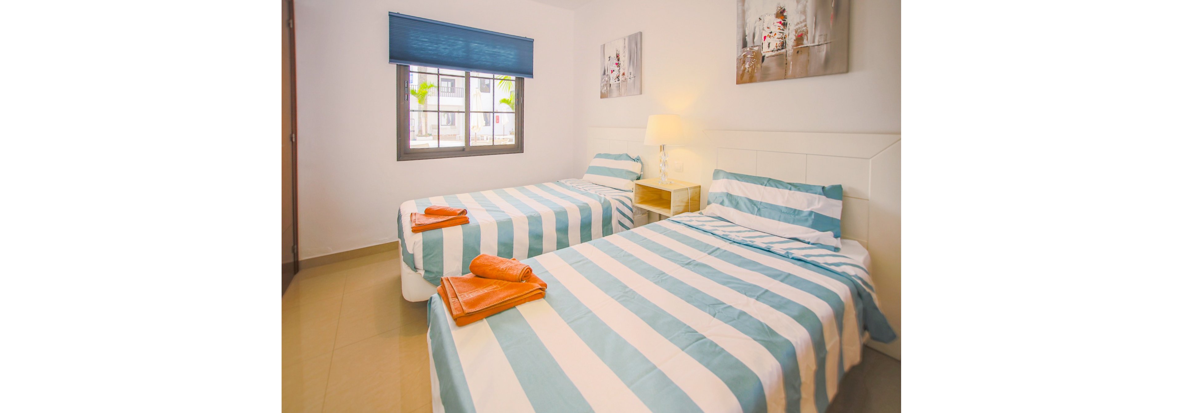 A clean, modern bedroom features two single beds with blue and white striped bedding. The room is softly lit by a table lamp on a small nightstand between the beds. A window with a blue blind lets in natural light and shows a glimpse of buildings and palm trees outside. Simple wall art and neatly folded orange towels on each bed add color accents to the neutral space.