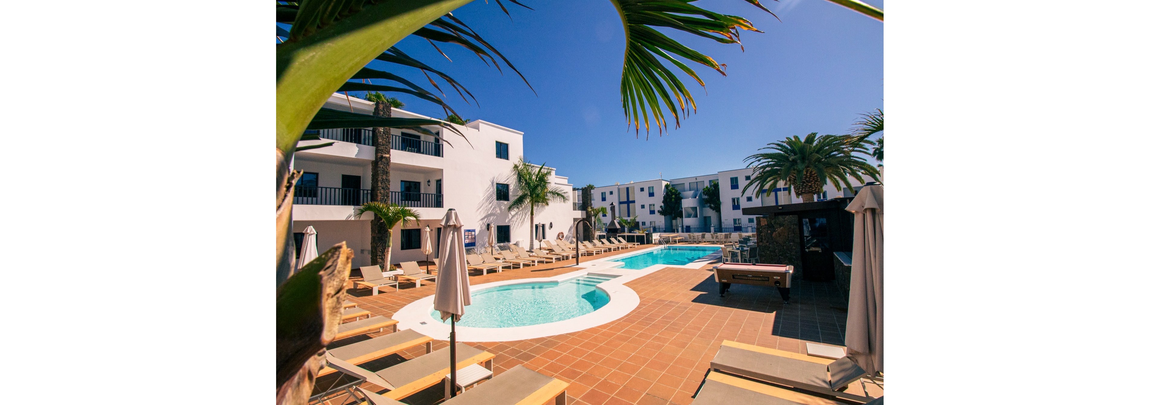A bright resort courtyard features a curved swimming pool surrounded by sun loungers and umbrellas. White modern apartment-style buildings frame the pool area under a clear blue sky. Palm trees and tropical plants add a vacation atmosphere to the scene. The overall setting suggests a warm, relaxing holiday destination with ample outdoor seating.