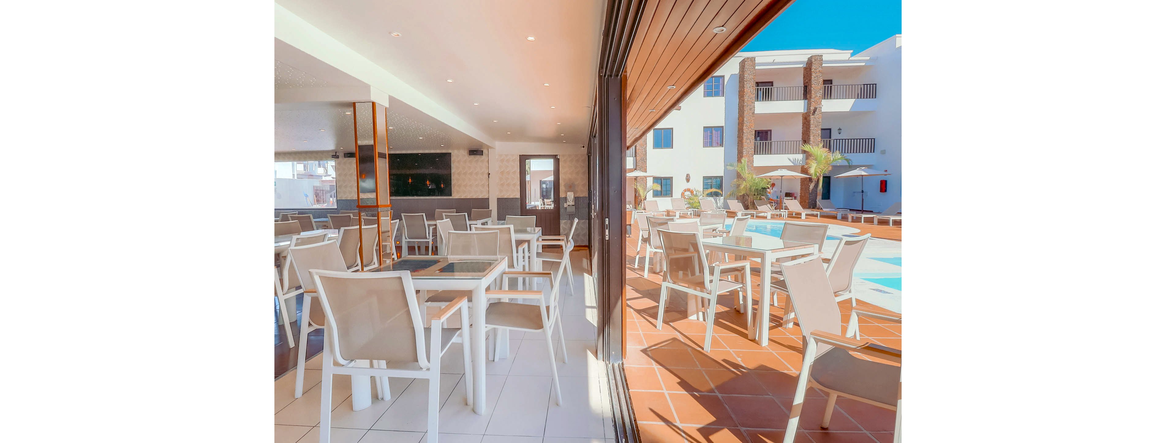Spacious indoor and outdoor dining areas are shown at a modern resort. White tables and chairs line both the interior tiled floor and the sunny poolside terrace. Large sliding glass doors connect the two spaces, creating an open, airy atmosphere. The scene is brightly lit with clear skies, emphasizing relaxation and vacation leisure.