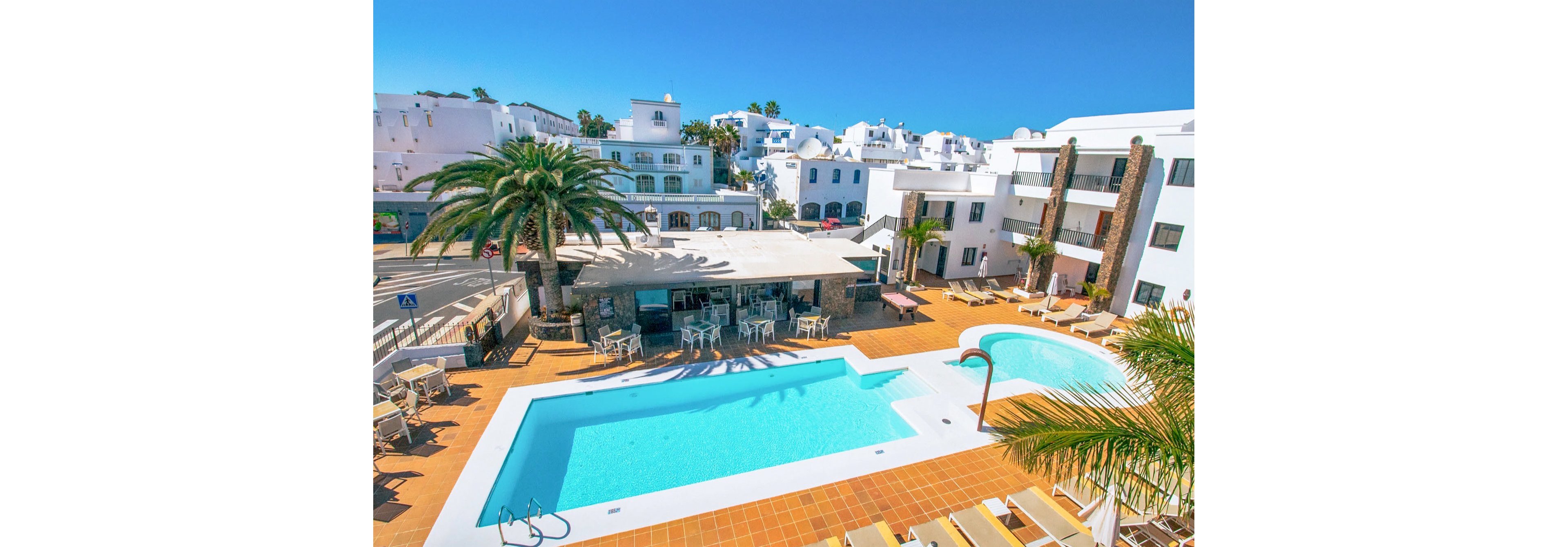 An outdoor resort courtyard features a bright blue swimming pool surrounded by sun loungers and palm trees. White Mediterranean-style buildings with balconies and terraces frame the pool area. A smaller curved pool or children’s pool sits beside the main rectangular pool. Tables and chairs are arranged under a shaded patio near the pool, suggesting a bar or café area. The clear blue sky and vivid colors create a warm, inviting holiday atmosphere.