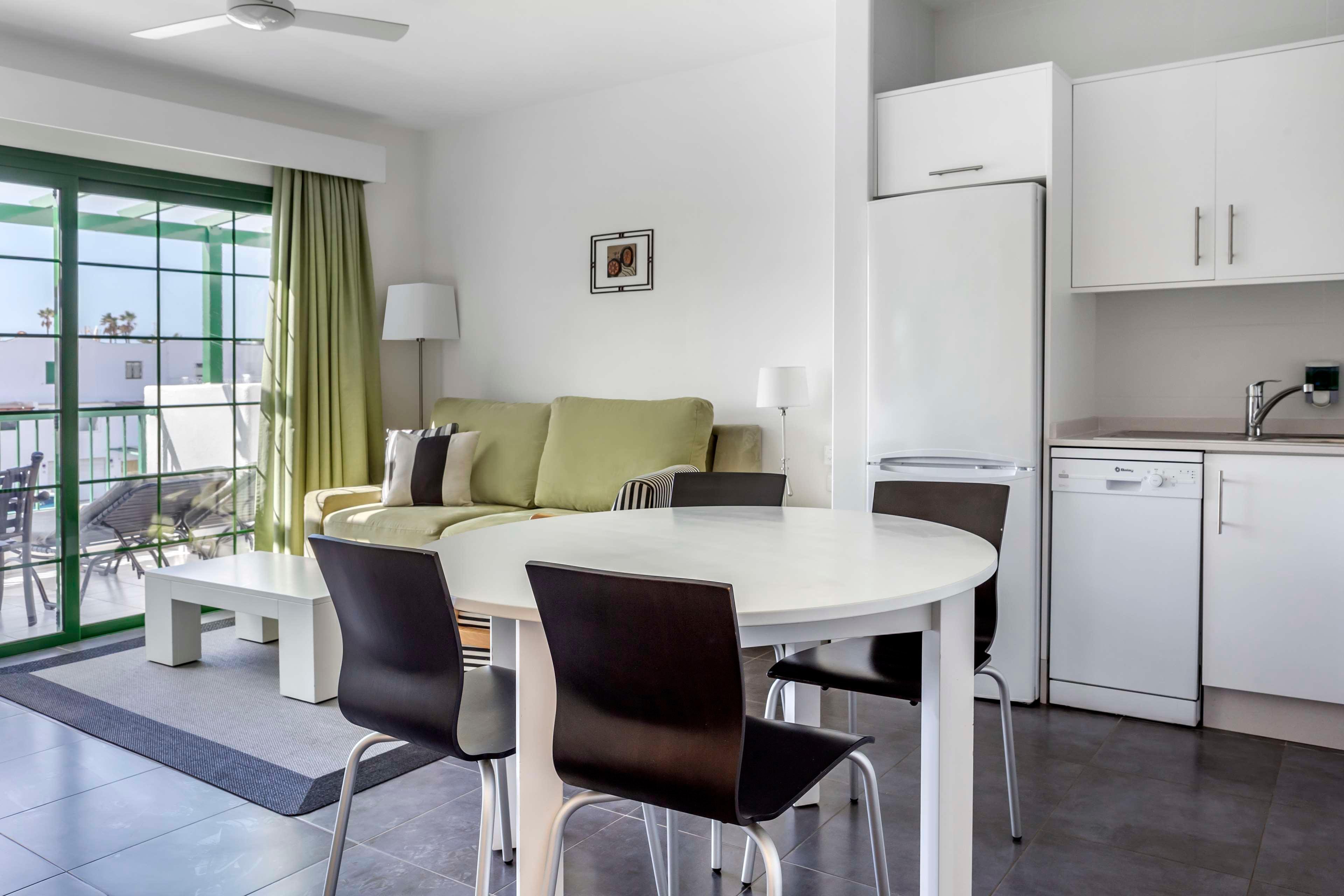 Contemporary open-plan apartment interior featuring a small dining table with dark chairs in the foreground. A light green sofa with cushions and a coffee table sit near large sliding glass doors leading to a balcony. The compact kitchen area includes white cabinets, a tall refrigerator, and a dishwasher. Neutral tones with green accents and ample natural light create a clean, airy atmosphere.