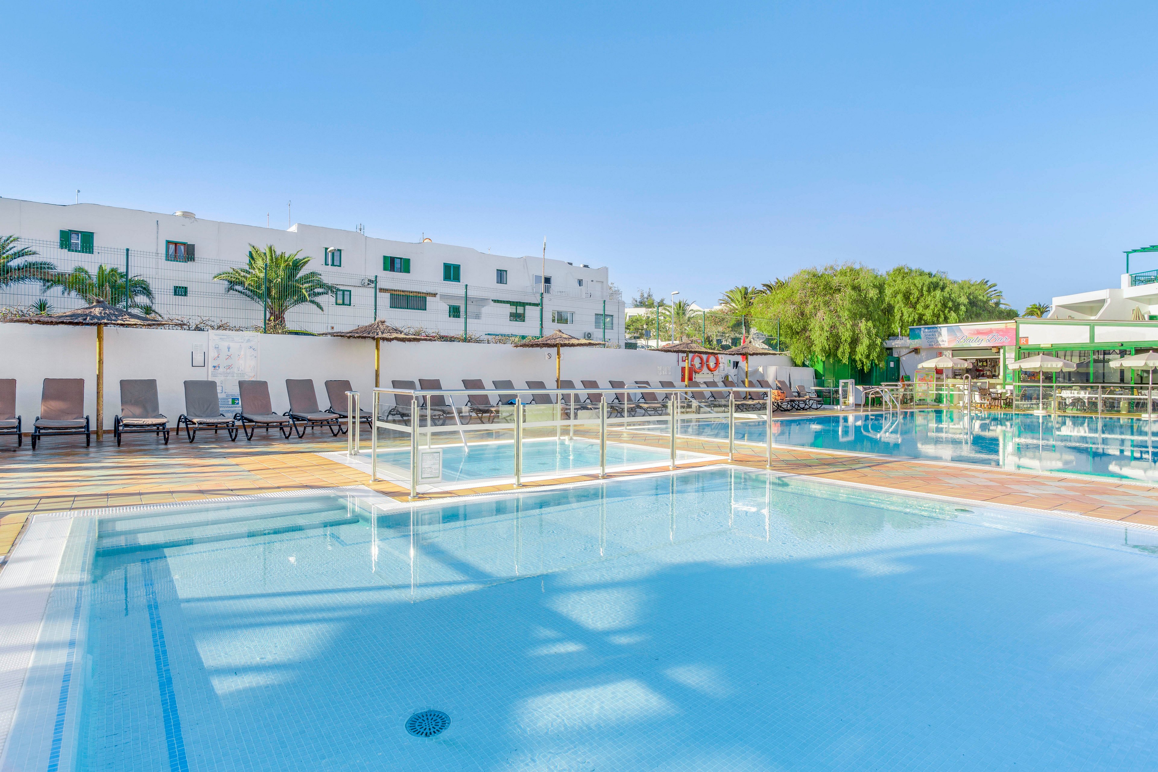 An outdoor swimming pool is shown under a clear blue sky at a resort. Sun loungers line the poolside deck, facing the water and ready for guests. A white multi-story building with green window shutters stands in the background, along with palm trees and lush greenery. The calm, reflective water and bright sunlight create a relaxing vacation atmosphere.