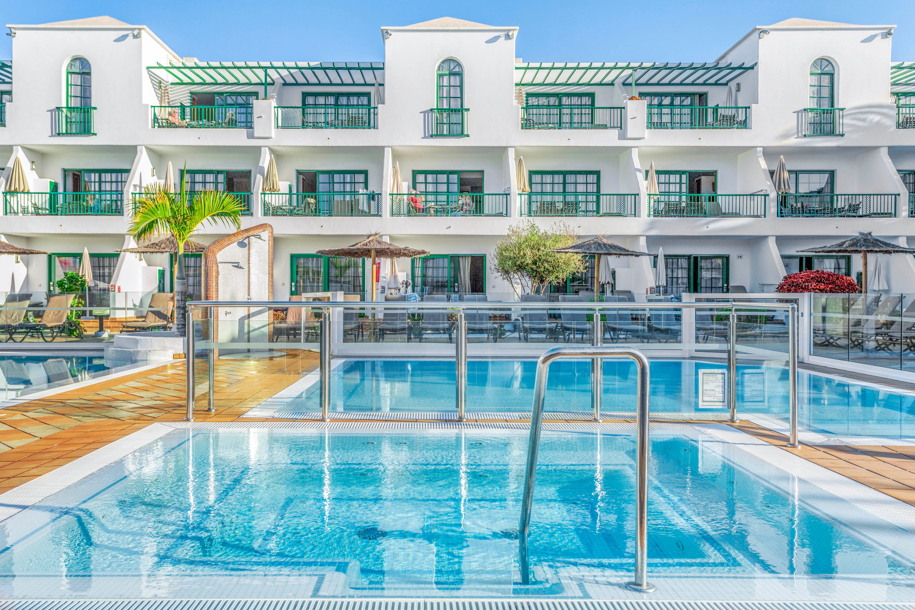 A clear blue swimming pool sits in the foreground of a bright resort complex. Behind the pool, white multi-story apartments with green railings and balconies face the water. Sun loungers, umbrellas, and potted plants line the poolside deck, suggesting a relaxing holiday setting. The sky is clear and blue, enhancing the fresh, sunny atmosphere of the scene.