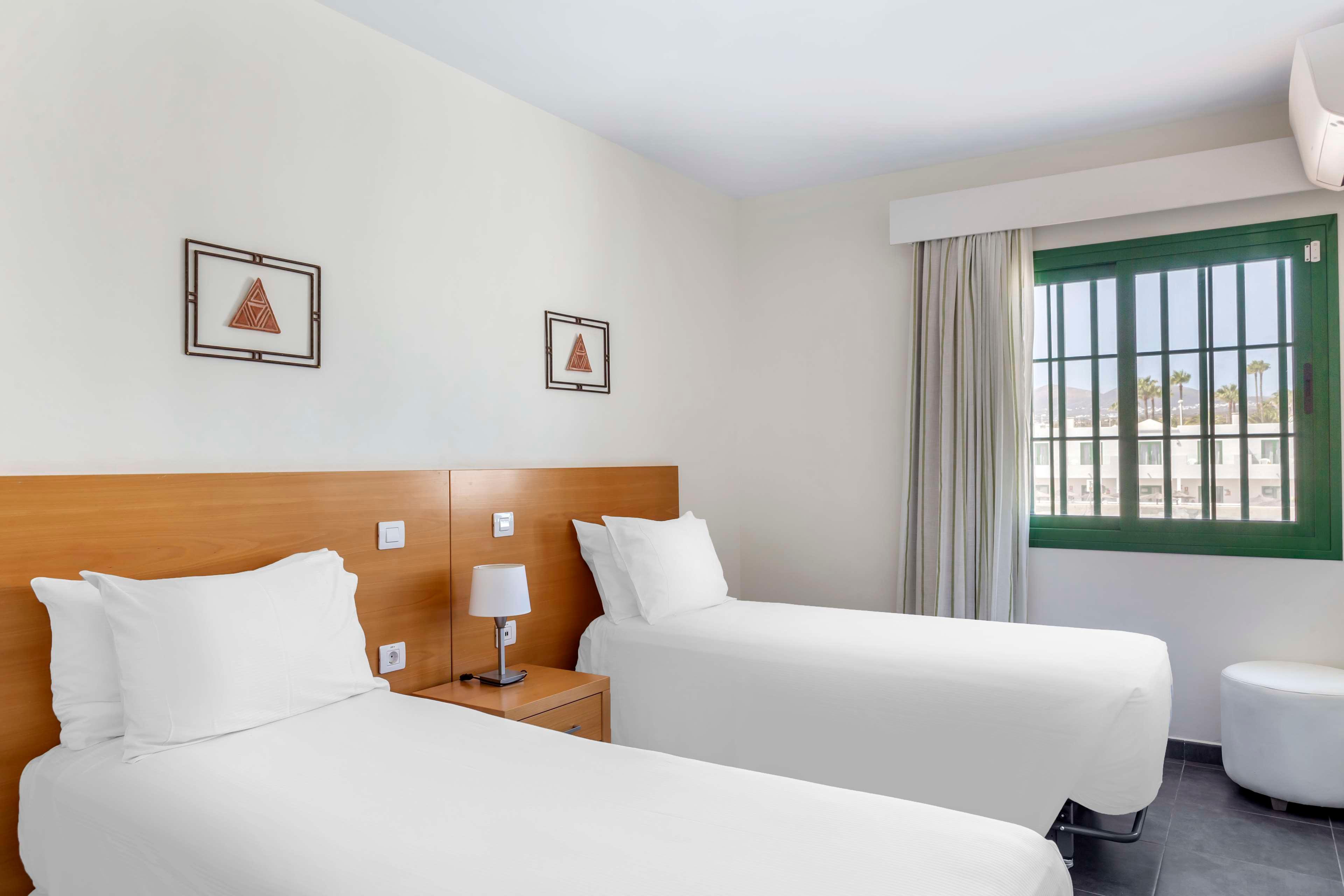 A clean, modern hotel room features two neatly made twin beds with white linens and wooden headboards. A small bedside table with a lamp and telephone sits between the beds. Minimalist artwork hangs on the wall above the headboards, and a large green-framed window lets in natural light. Neutral walls and curtains create a bright, simple, and inviting atmosphere.