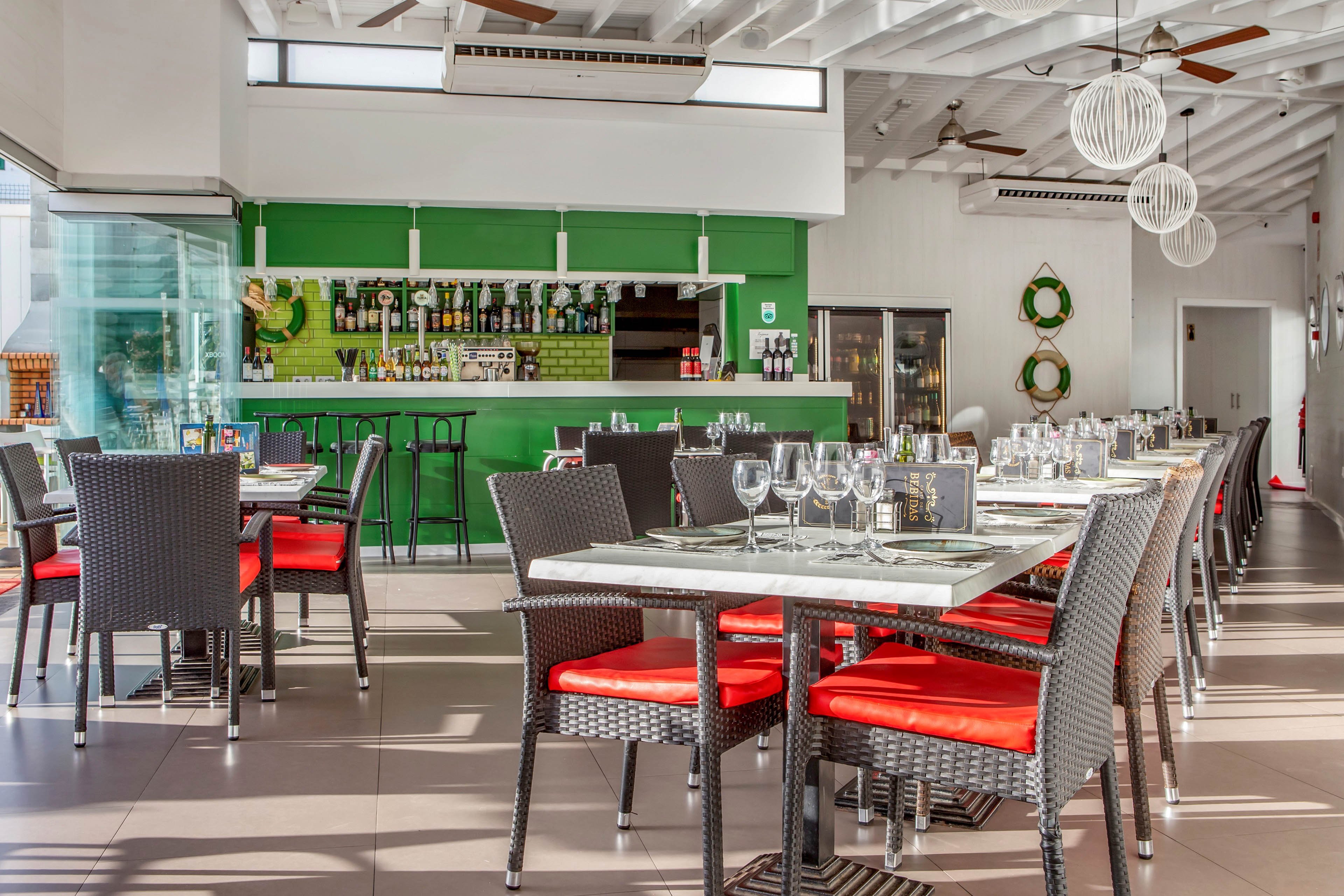 Contemporary restaurant dining area with neatly arranged tables and chairs is shown in bright natural light. The space features a green bar wall stocked with bottles and glassware in the background. Black wicker chairs with vivid red cushions surround marble-topped tables set with wine glasses, menus, and cutlery. Clean lines, white ceiling beams, and decorative hanging lights create a fresh, inviting atmosphere.