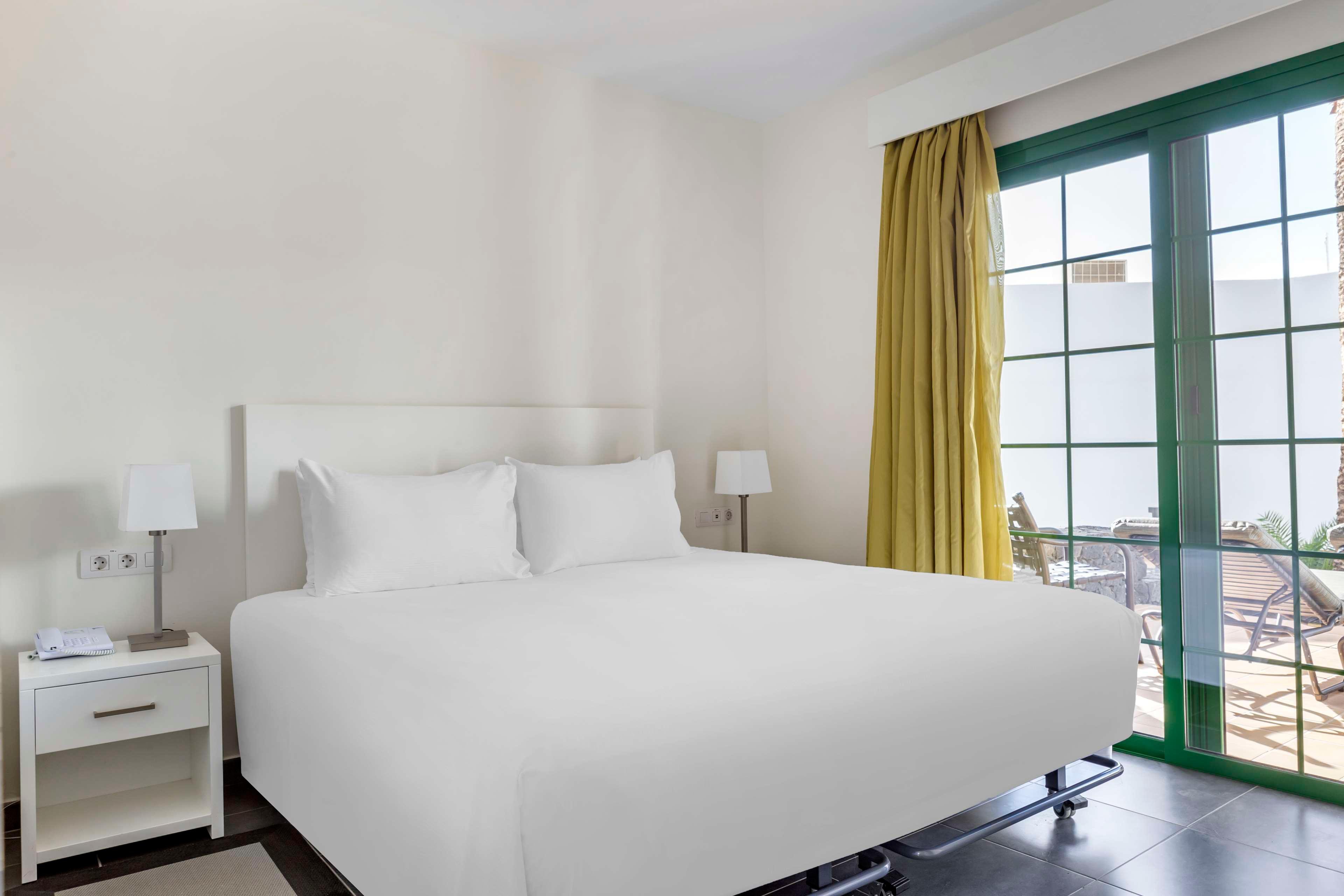 A neatly made double bed with white linens is centered in a bright hotel bedroom. Two bedside tables with matching lamps flank the bed, emphasizing a clean and minimal design. Large green-framed sliding glass doors open to an outdoor terrace with chairs and a table. Yellow curtains and neutral walls create a calm, inviting atmosphere suitable for relaxation or vacation stays.