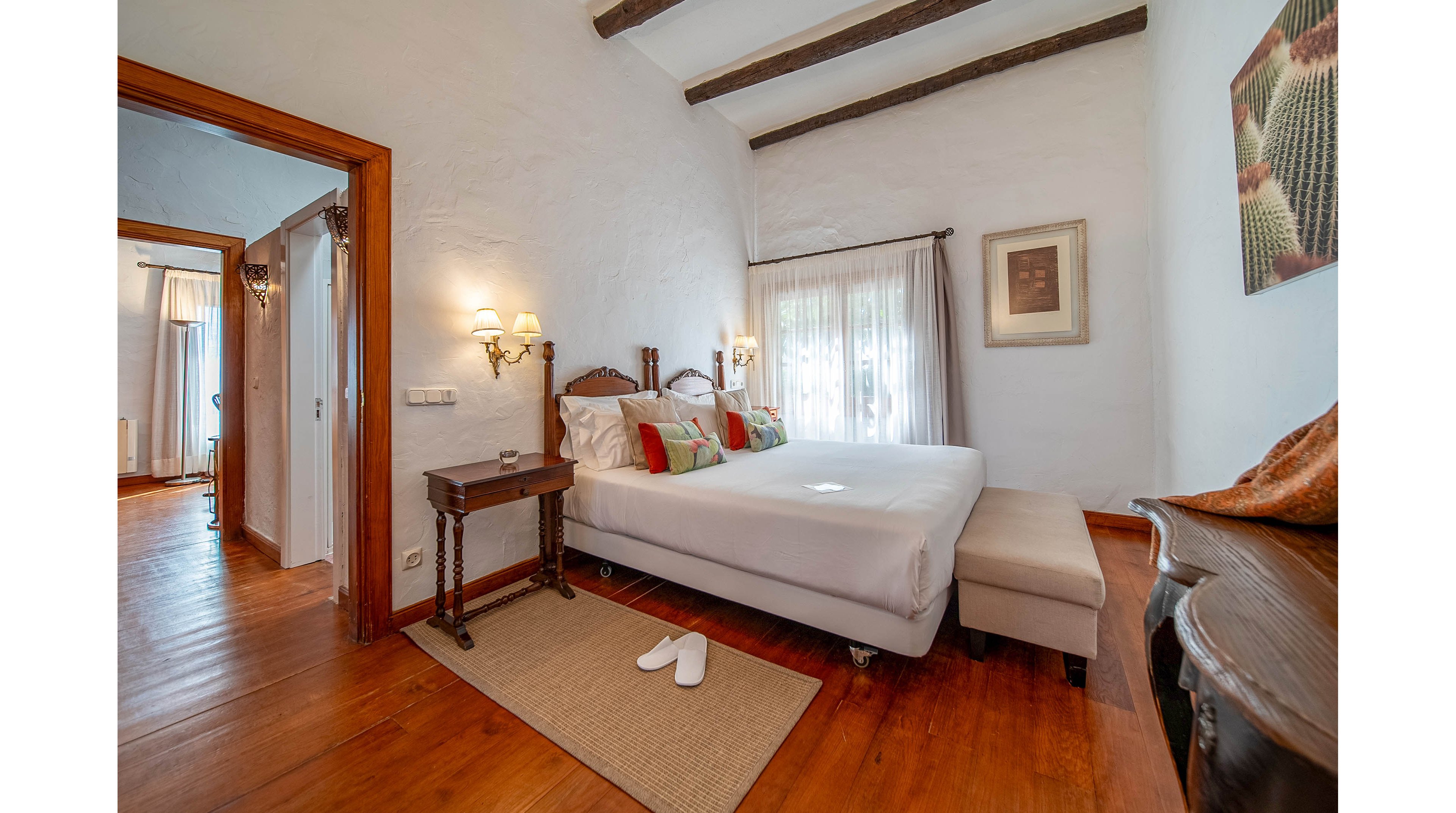A cozy bedroom features a neatly made double bed with white linens and colorful accent pillows. The room has warm wooden floors, exposed ceiling beams, and soft natural light coming through sheer curtains. Classic wall sconces, a small bedside table, and a cushioned bench at the foot of the bed add comfort. An open doorway reveals an adjoining room, enhancing the spacious and inviting feel of the interior.