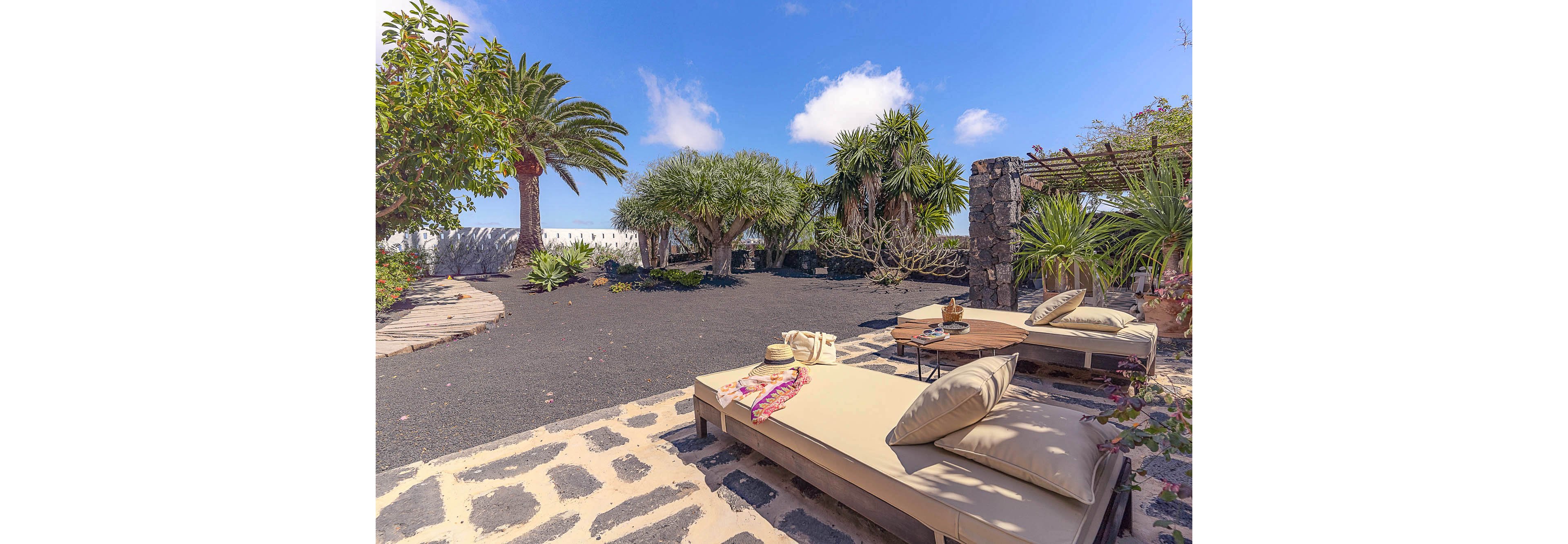 Two cushioned lounge chairs are set on a stone patio, facing a lush tropical garden with palm trees and volcanic black gravel. The scene is bathed in bright sunlight under a blue sky with scattered clouds. A straw hat, scarf, and tray with drinks add a relaxed, vacation-like atmosphere. The setting is tranquil and inviting, ideal for leisure and outdoor relaxation.