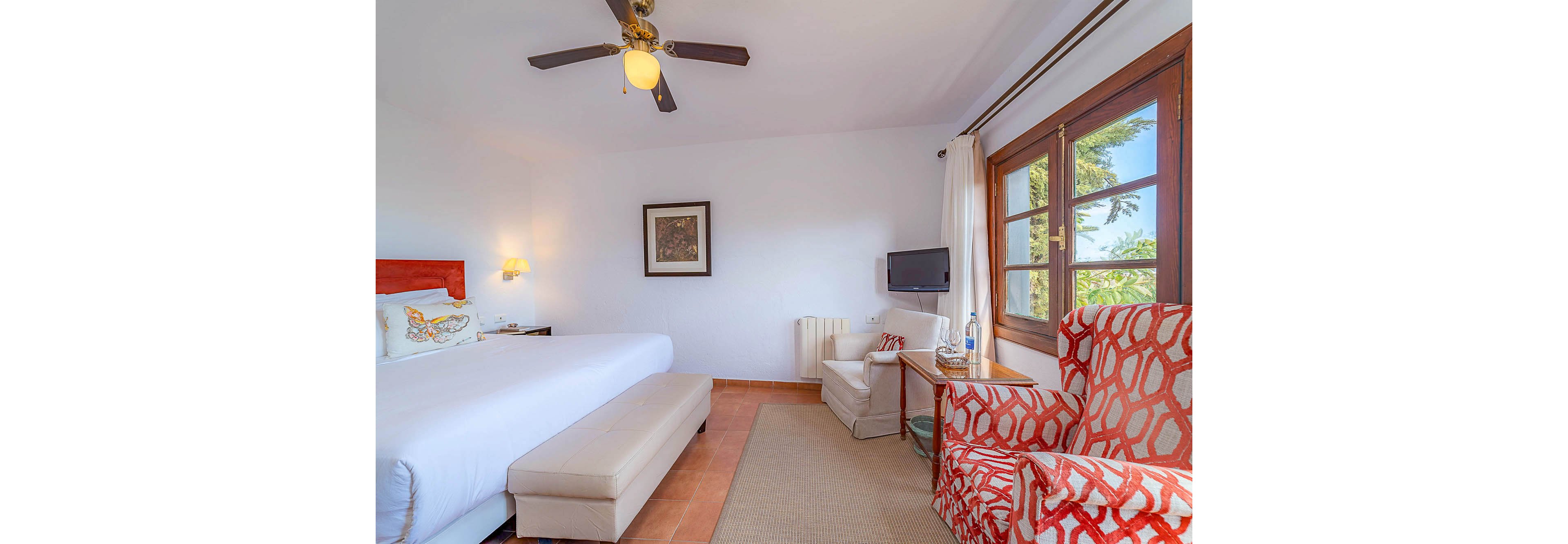 A spacious hotel room features a large bed with white linens and a cushioned bench at the foot. The setting includes a patterned orange armchair, a small television, and a window letting in natural light. The decor is modern with a neutral palette, accented by pops of color from the chair and pillows. The room appears inviting and comfortable, ideal for relaxation.