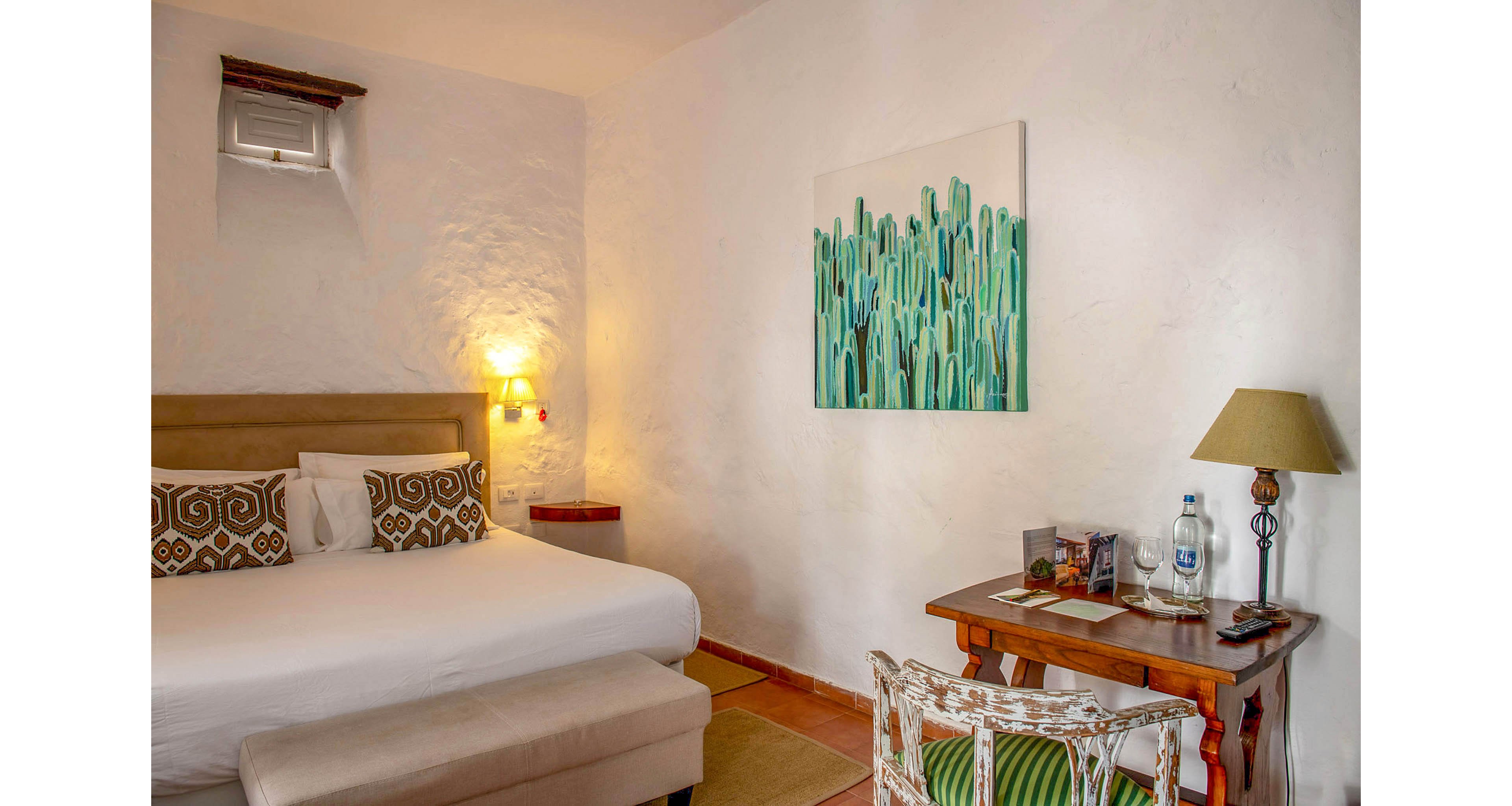 A comfortable hotel room features a large bed with patterned pillows and a cushioned bench at the foot. The setting includes a wooden desk with a lamp, bottled water, and reading materials, while a green cactus-themed painting decorates the white textured wall. Warm lighting and neutral tones create a relaxing, inviting atmosphere.