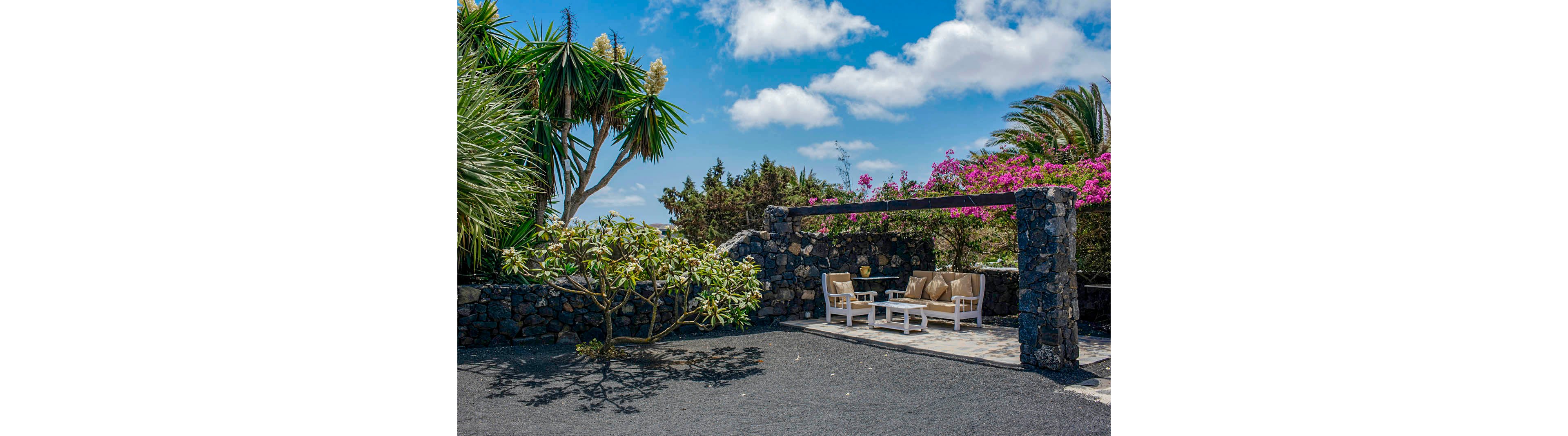 A cozy outdoor seating area features cushioned chairs and a table under a pergola, surrounded by lush tropical plants and a stone wall. The setting is sunlit with a bright blue sky and scattered clouds overhead. Vibrant greenery and blooming flowers add color to the tranquil patio scene. The ground is covered with dark gravel, enhancing the natural ambiance. No visible text or numbers are present in the image.