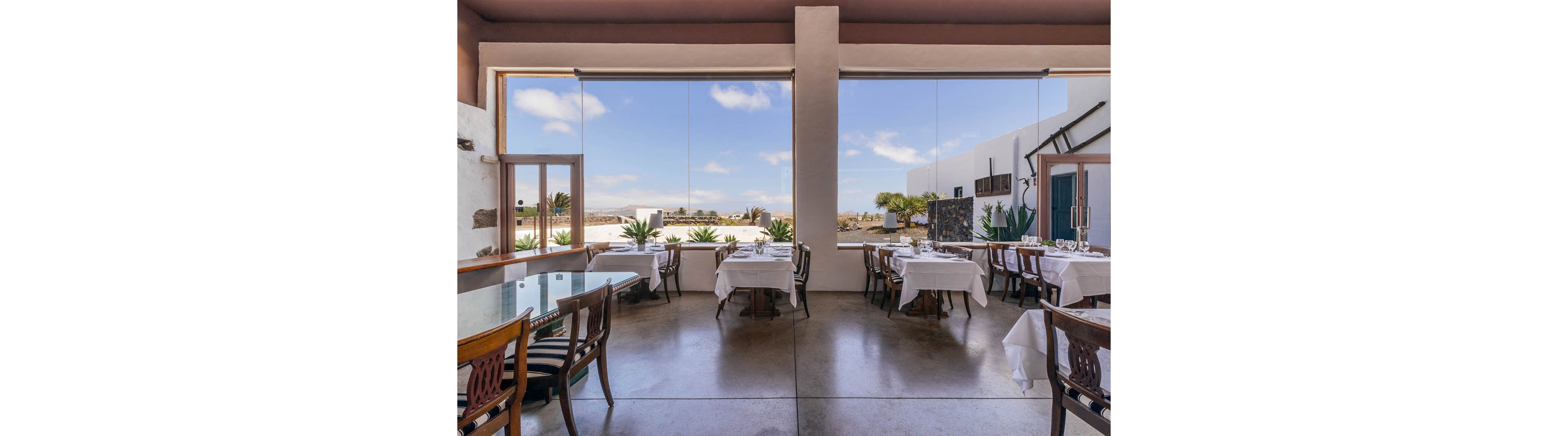 A contemporary restaurant dining area features large floor-to-ceiling windows overlooking a bright outdoor landscape. Tables are set with white tablecloths and glassware, creating an elegant and inviting atmosphere. The space is filled with natural light, highlighting minimalist decor and polished concrete floors. No visible text or numbers are present in the image.