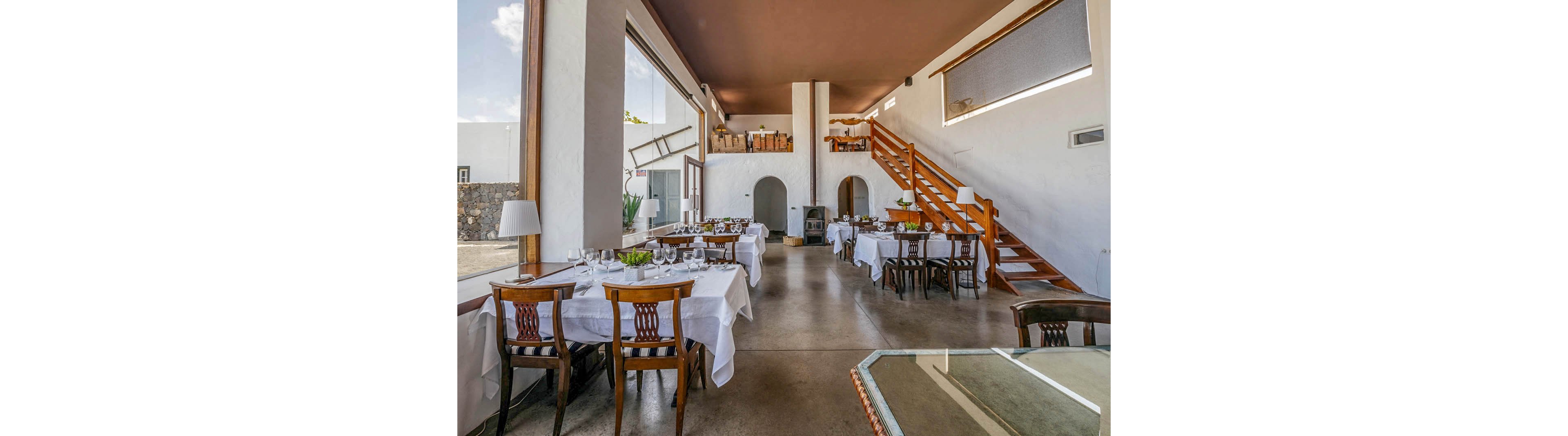 Spacious restaurant dining area featuring large windows and natural light. Tables are set with white linens, glassware, and wooden chairs, creating an inviting atmosphere. The room includes a wooden staircase leading to an upper level, with minimalist decor and neutral tones throughout. No visible people or text are present in the scene.