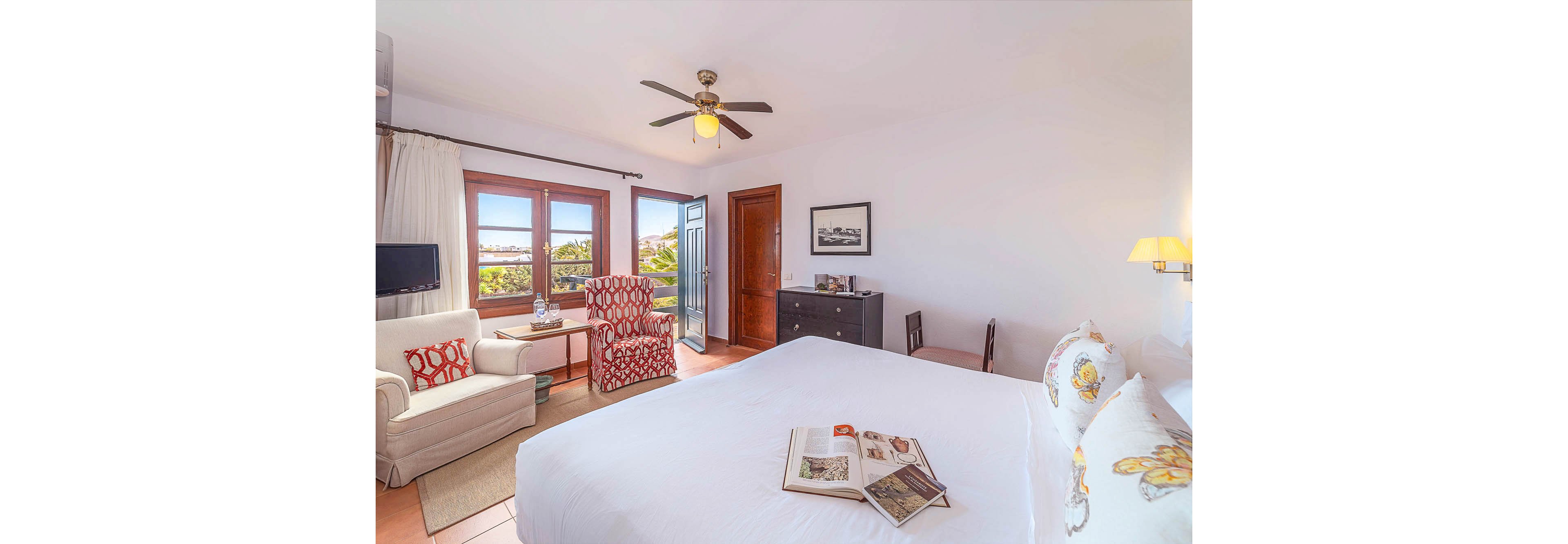 A spacious hotel room features a large bed with floral pillows, a comfortable armchair, and a small sofa arranged near a window and open balcony door. The room is decorated in warm, neutral tones with pops of red and yellow, and includes a ceiling fan, wall-mounted TV, and dresser. Natural light fills the space, highlighting the inviting and relaxed atmosphere, while magazines rest on the bed.