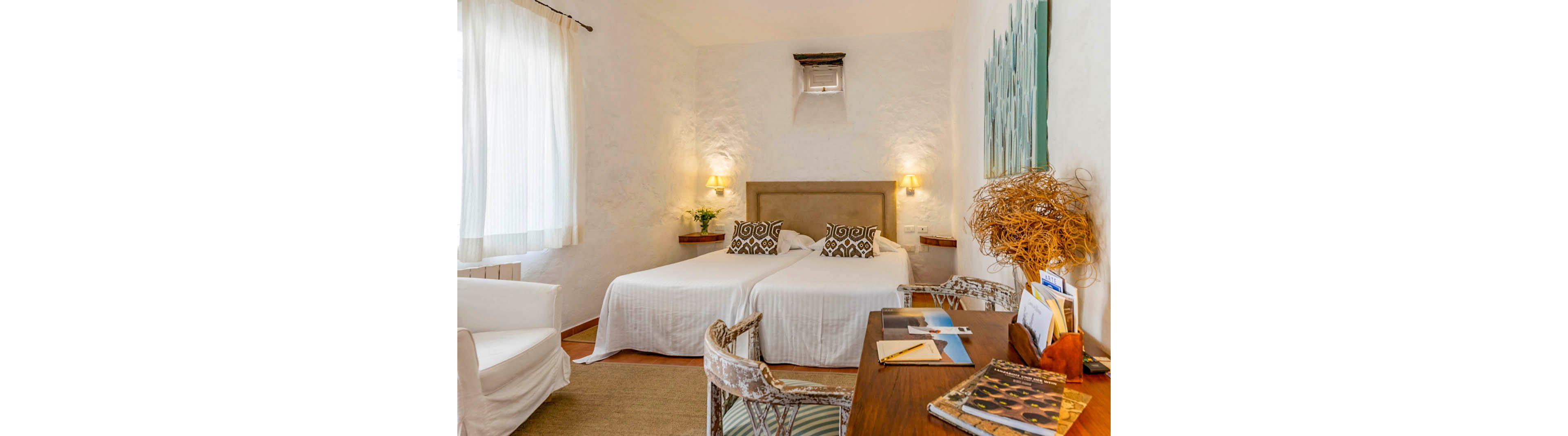 A well-lit hotel room features two twin beds with white linens and patterned pillows, set against a textured white wall. The space includes a wooden desk with books and papers, wicker chairs, and a comfortable armchair near a large window with sheer curtains. Warm lighting, natural decor elements, and neutral tones create a welcoming and serene atmosphere.