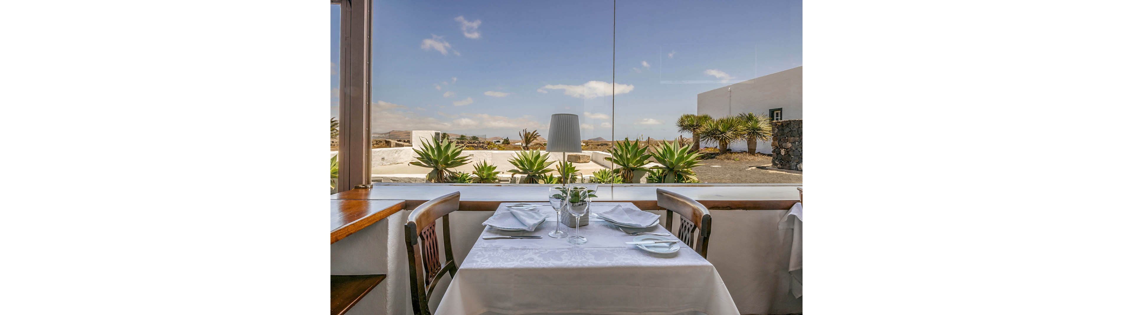 A neatly set dining table for two is positioned by a large window, offering a clear view of a sunlit outdoor landscape with palm plants and white buildings. The table features white linens, glassware, and folded napkins, creating a refined and inviting atmosphere. The scene is bright and tranquil, with natural light enhancing the clean, minimalist decor.