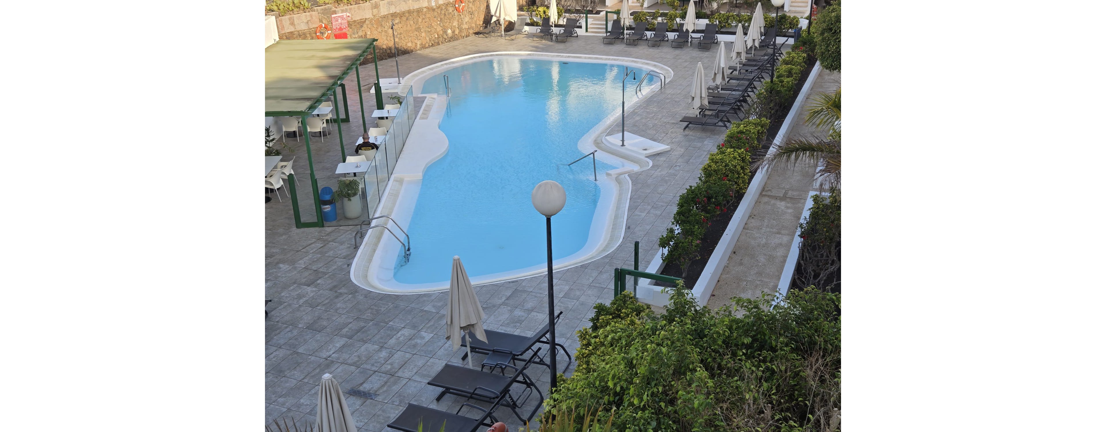 An outdoor swimming pool is surrounded by a tiled deck at a hotel or resort. Numerous sun loungers and closed umbrellas line the poolside, with no people present. A covered seating area and glass safety barriers are visible on one side, while green shrubs and a stone wall frame the space. The scene suggests a quiet, early or off-season moment in a leisure setting.