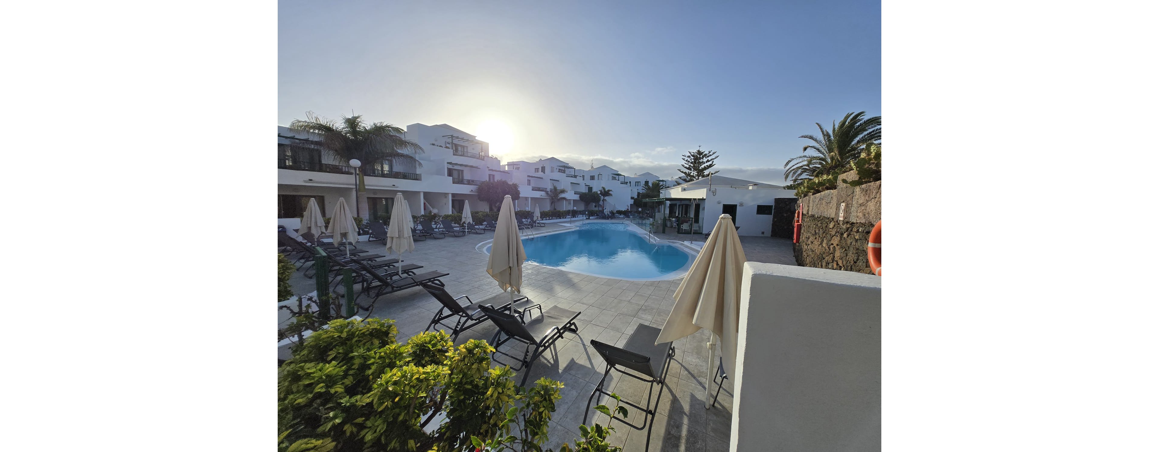 A curved outdoor swimming pool is surrounded by sun loungers and umbrellas in a modern resort complex. White multi-story buildings frame the pool area, with balconies overlooking the scene. The early morning or late afternoon sun creates long shadows and a warm glow across the tiled terrace. Green plants and palm trees add a tropical touch to the tranquil vacation setting.