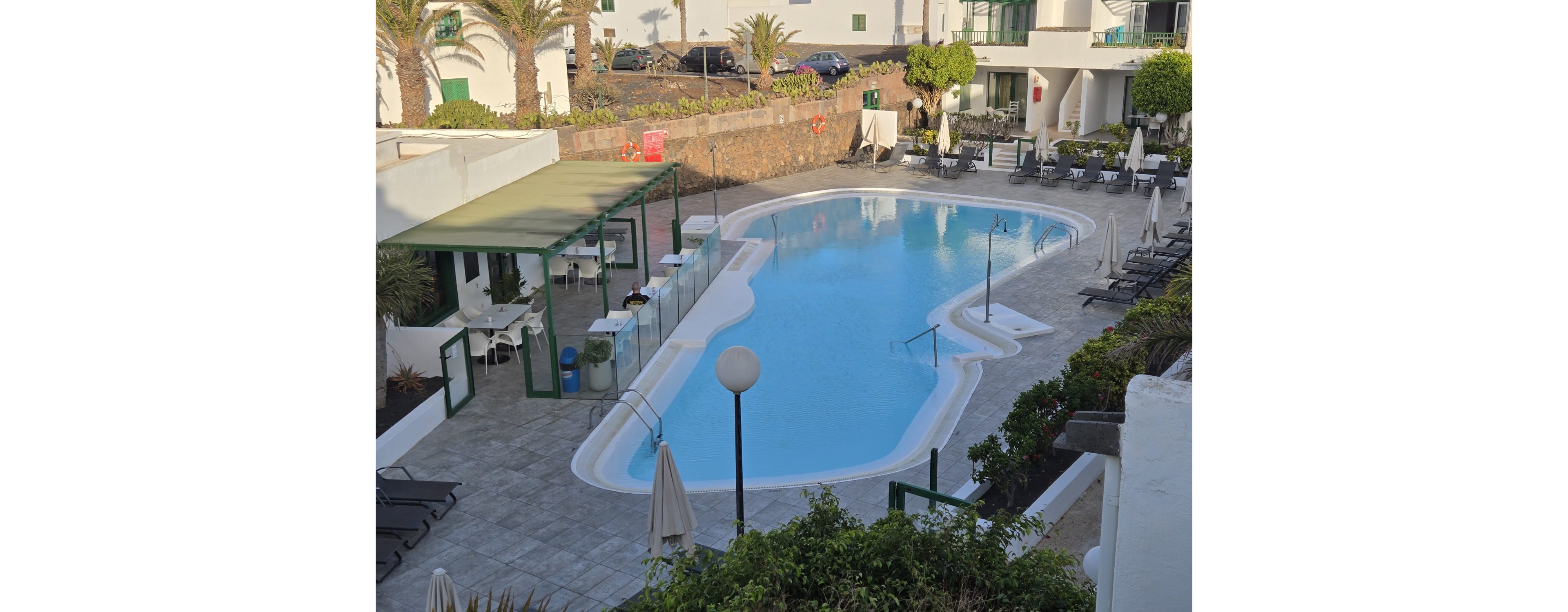 An empty outdoor swimming pool sits in the center of a hotel courtyard. Sun loungers and closed umbrellas line the tiled deck around the water, suggesting a quiet moment between uses. A small poolside bar or café with tables and chairs is visible to one side, enclosed by glass fencing. White buildings, potted greenery, and palm trees surround the area, creating a relaxed vacation atmosphere.
