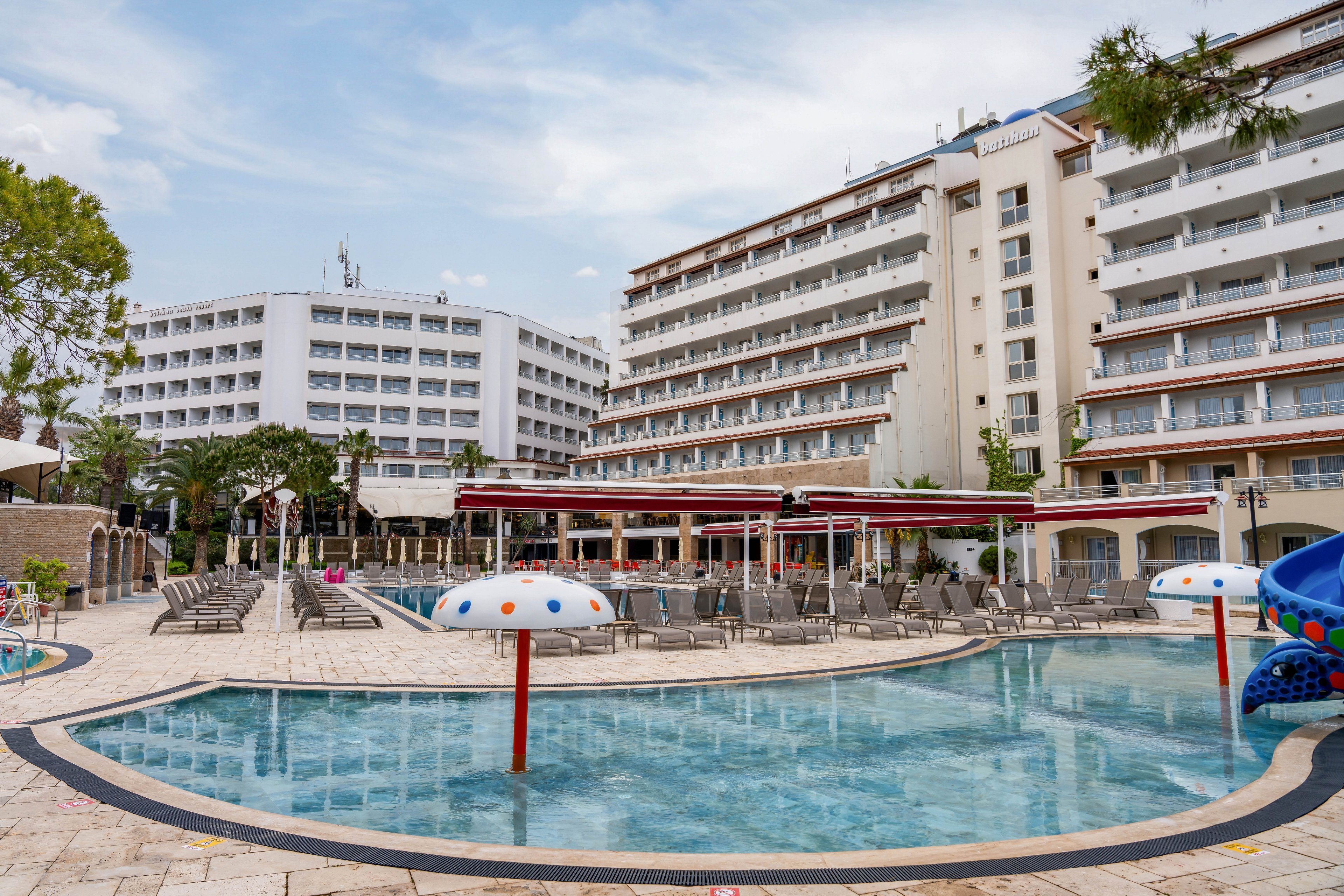 A large multi-story resort hotel overlooks an expansive outdoor pool area. The foreground features a shallow pool with playful mushroom-shaped fountains and a small blue water slide. Numerous sun loungers and umbrellas are arranged neatly around the pool deck, suggesting a relaxing vacation setting. The sky is lightly overcast, giving the scene a calm and quiet atmosphere.