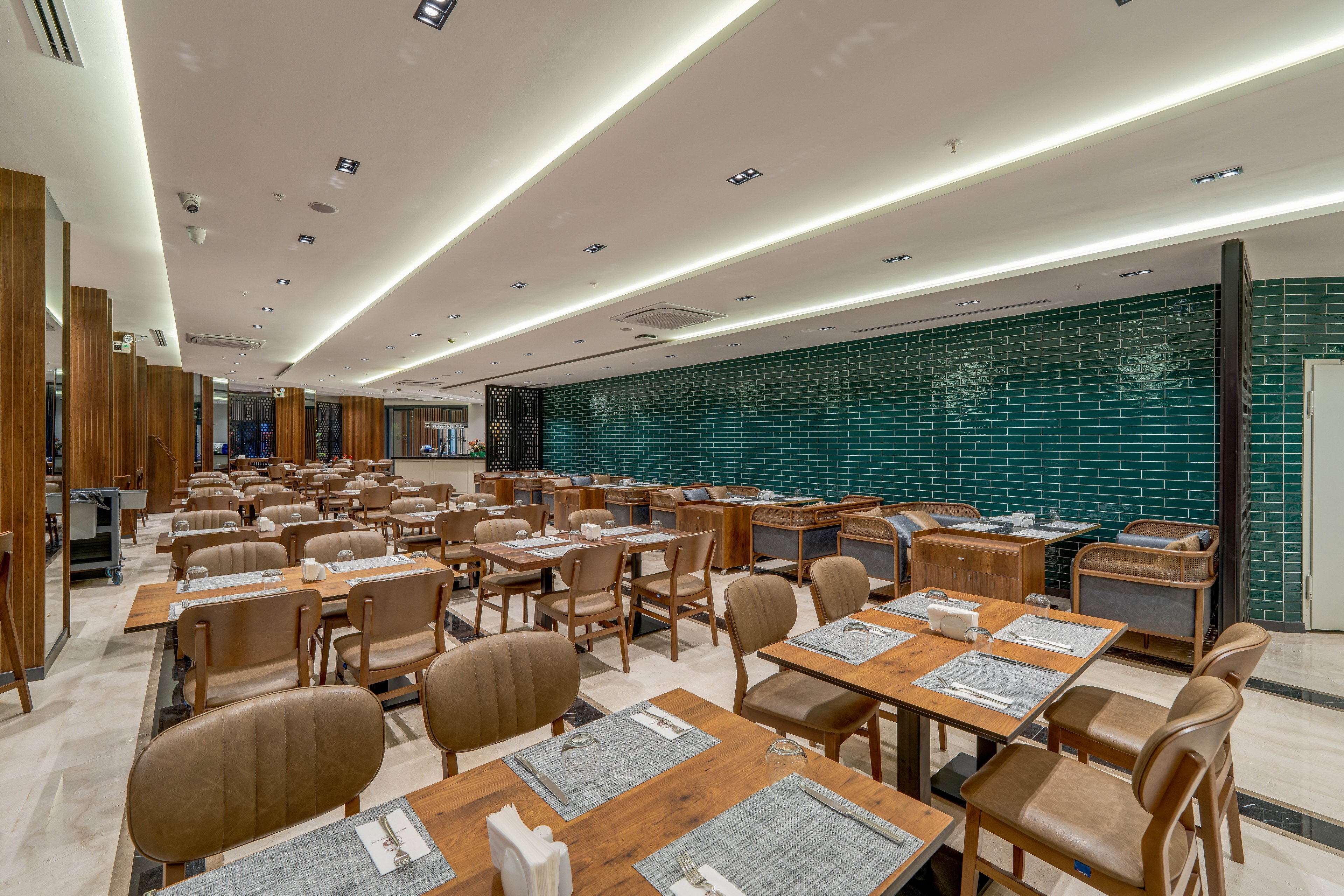 A contemporary restaurant dining area is shown with rows of wooden tables and cushioned chairs neatly arranged. The space is empty of people, emphasizing the clean and orderly setup. A long wall with glossy dark green tiles contrasts with the warm wood tones and beige flooring. Recessed ceiling lights and linear fixtures create bright, even illumination throughout the interior.