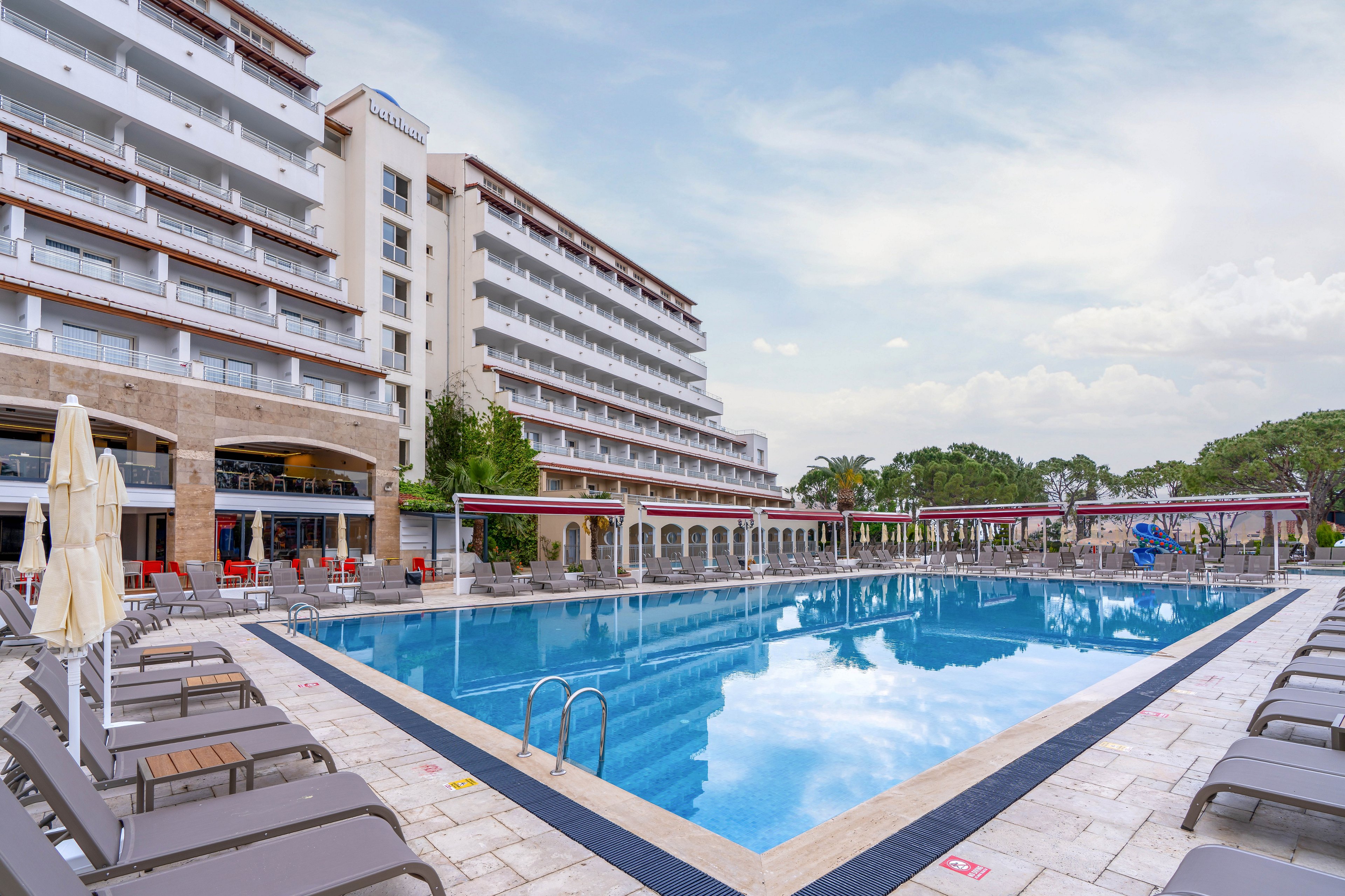A spacious outdoor swimming pool sits in front of a modern multi-story resort hotel. Rows of lounge chairs and umbrellas line the poolside deck, creating a relaxing vacation setting. The building features balconies overlooking the water and covered walkways along the ground floor. Trees and a small play area are visible in the background under a cloudy sky.