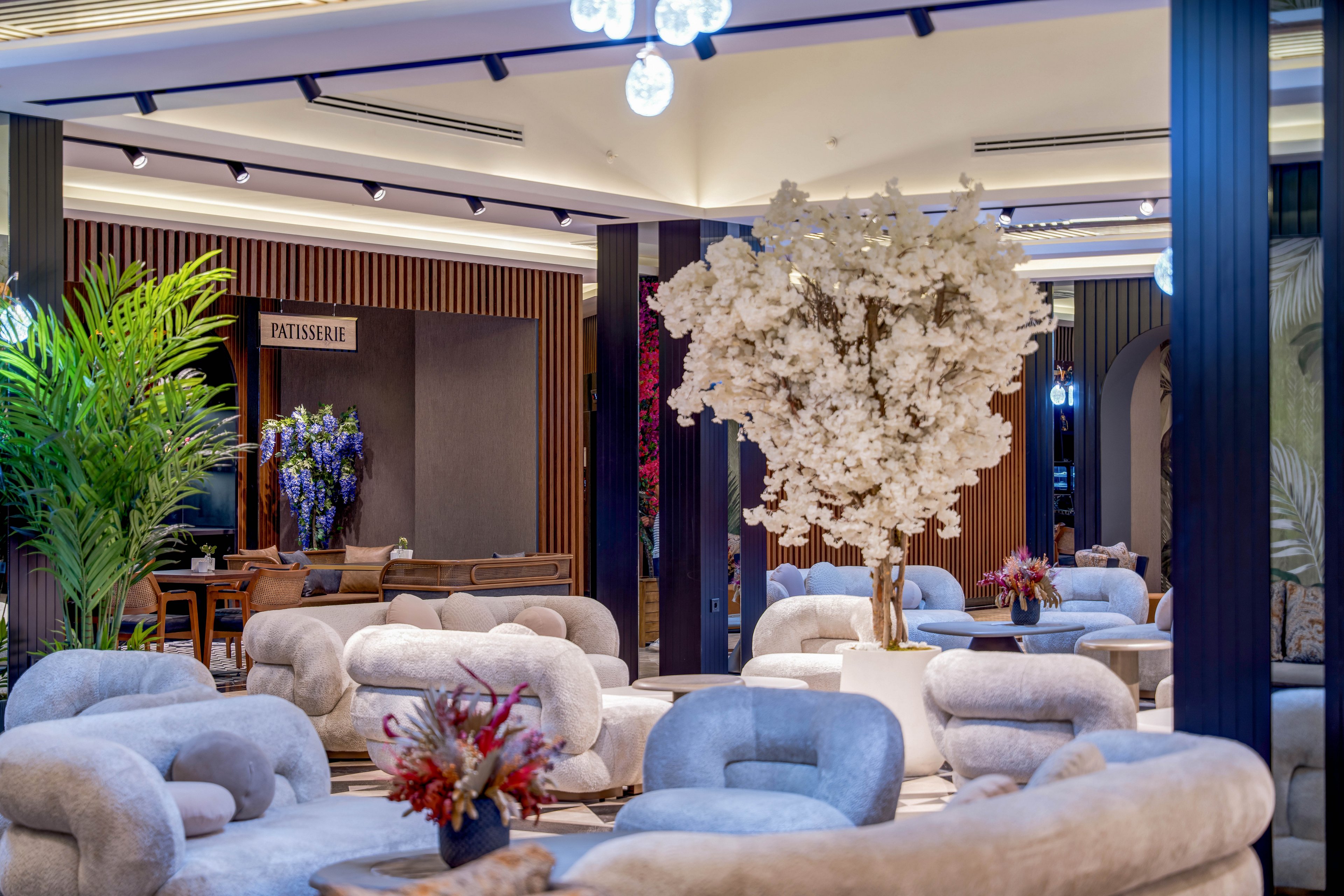 A contemporary lounge area features curved, plush sofas arranged around low tables. At the center stands a large white flowering tree in a decorative pot, creating a striking focal point. Warm ambient lighting, tall black columns, and wooden wall panels give the space a luxurious feel. In the background, a patisserie section and additional floral arrangements add color and depth to the interior scene.