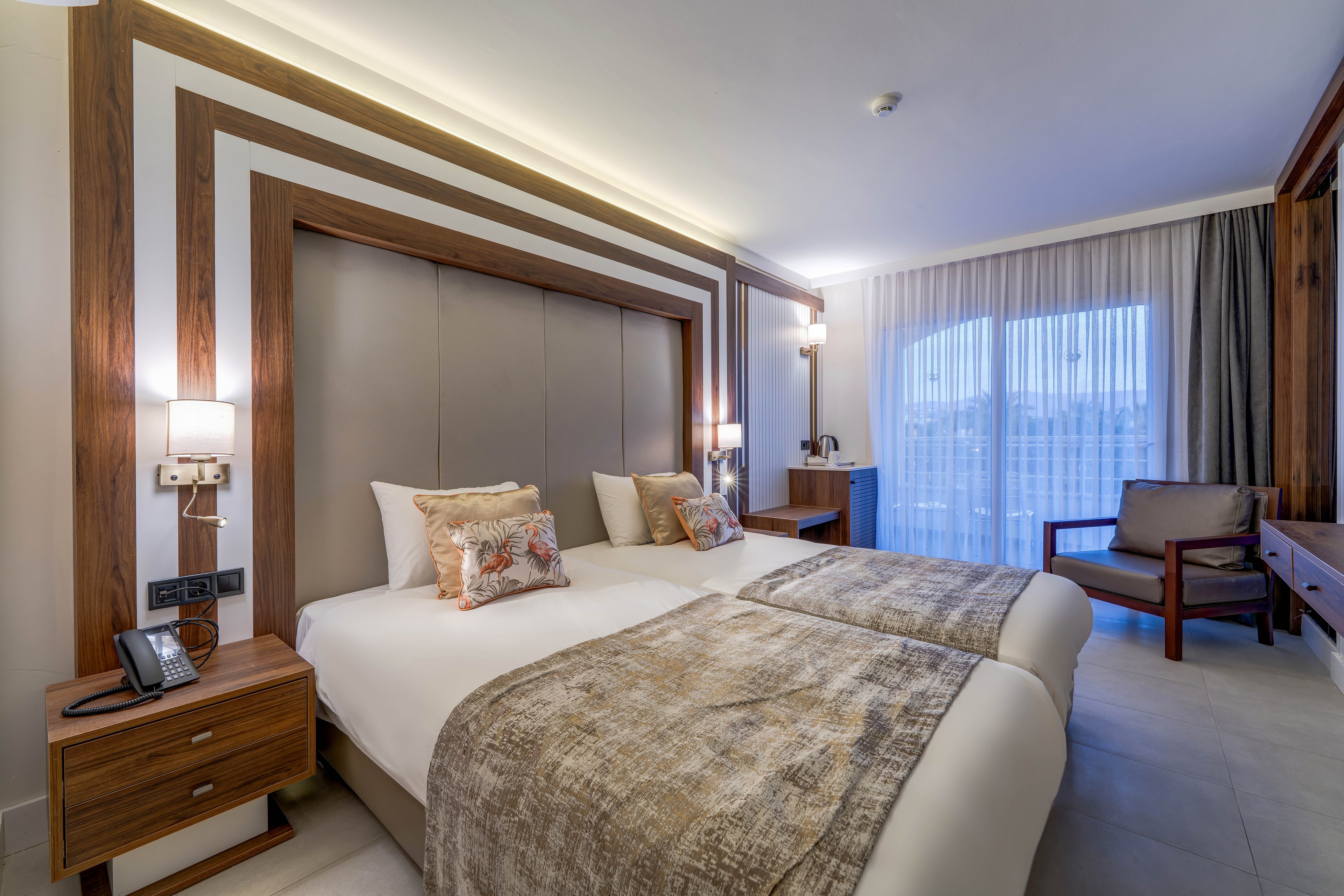 Contemporary hotel room featuring two neatly made twin beds with padded headboard and patterned throws. The space includes wooden bedside tables with lamps, a desk area, and a comfortable armchair. Large floor-to-ceiling windows with sheer curtains open to a balcony and outdoor view. Soft ambient lighting and neutral tones create a calm, inviting atmosphere suitable for relaxation or business travel.