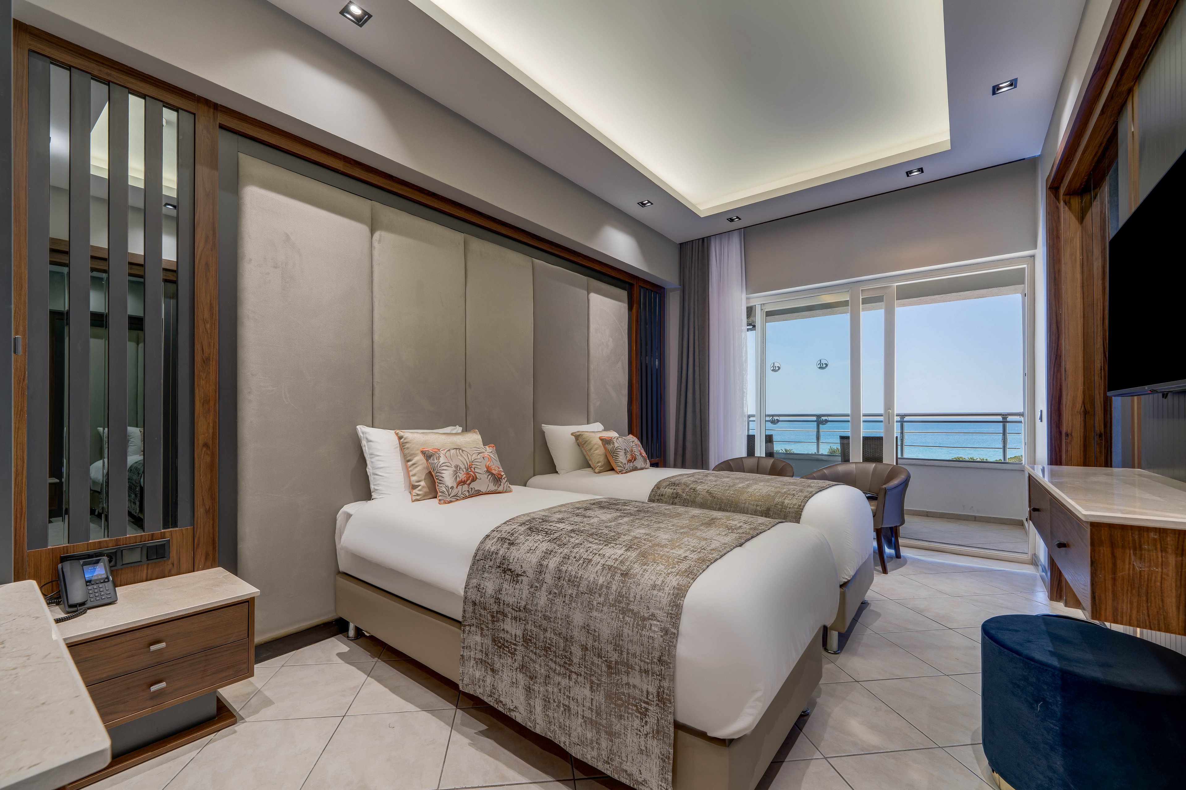 A contemporary hotel room features two neatly made twin beds with neutral bedding and decorative pillows. The space is designed with sleek wood accents, upholstered wall panels, and tiled flooring. A large window and glass door open to a balcony overlooking the sea and distant cable cars. Soft recessed ceiling lighting and bedside lamps create a calm, inviting atmosphere suitable for relaxation or business travel.