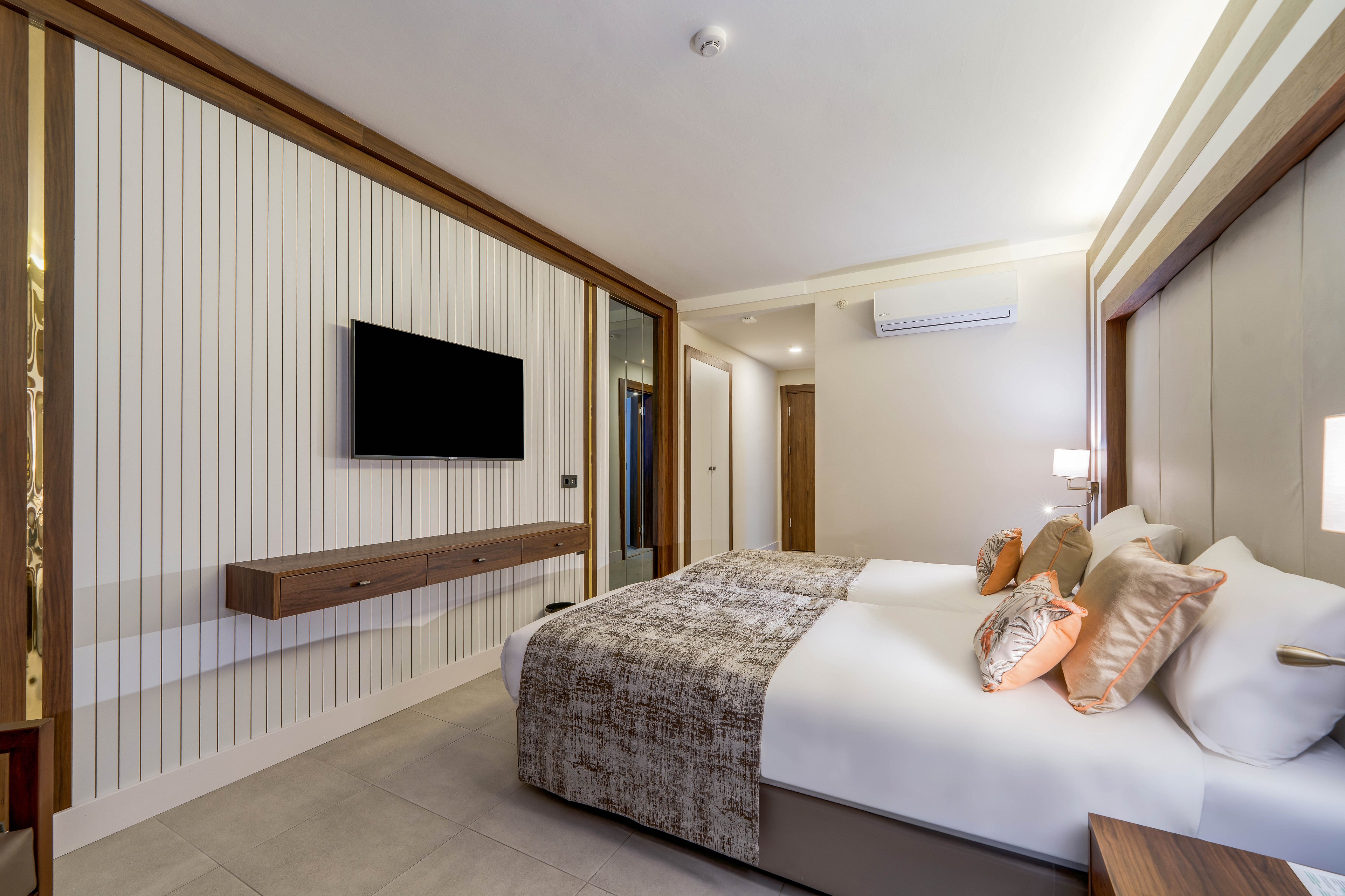 Contemporary hotel room featuring two neatly made twin beds with patterned throws and decorative pillows. A flat-screen television is mounted on a wall with vertical paneling above a floating wooden console. Warm lighting from bedside lamps and wall fixtures creates a cozy atmosphere. A doorway and mirrored wardrobe are visible, along with a wall-mounted air conditioning unit, emphasizing comfort and modern amenities.